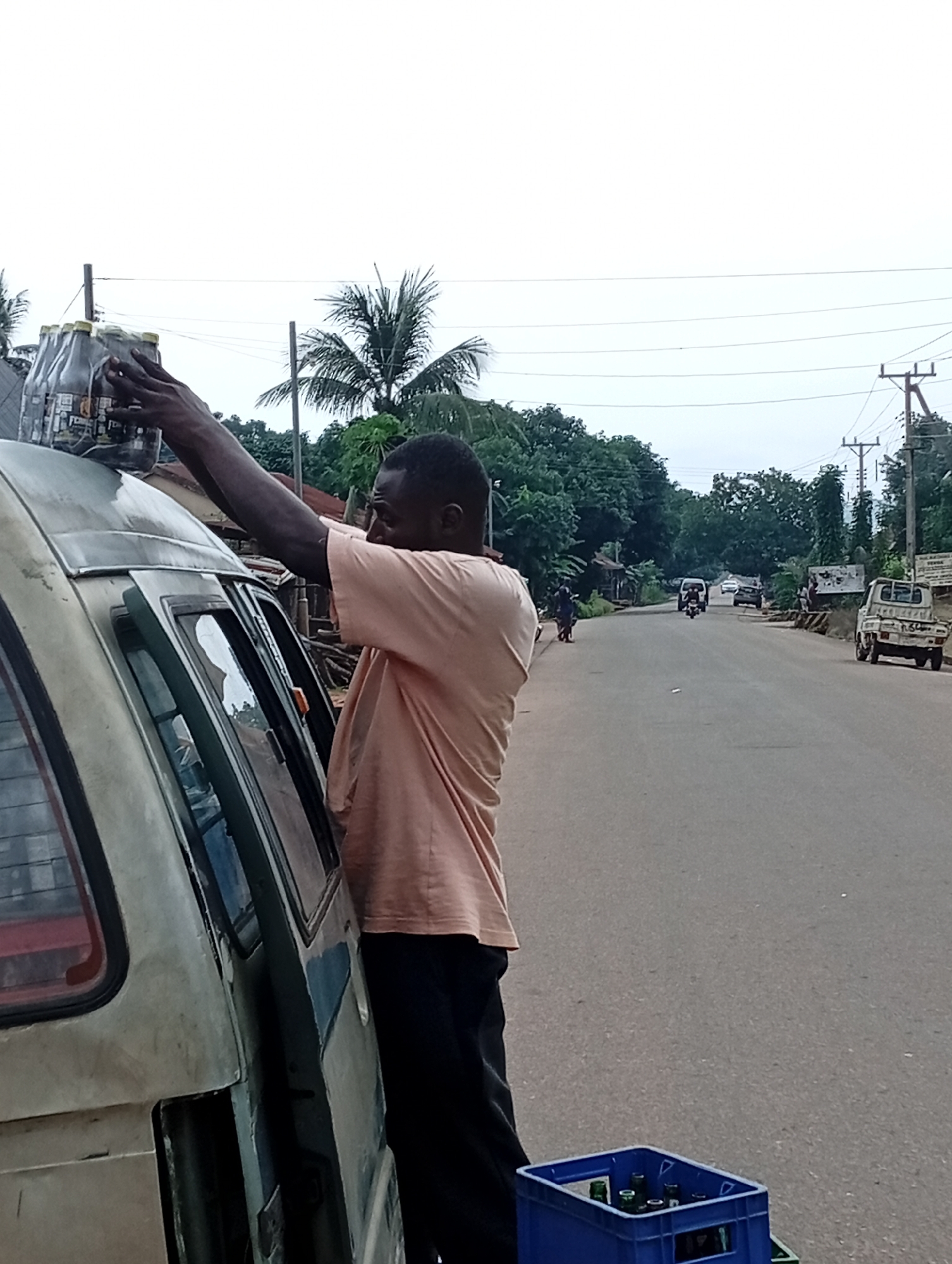 In Nsugbe, Nigeria, a Black man wearing a peach-colored t-shirt and dark pants is seen loading bottles of what appears to be alcoholic beverages onto the roof of a light-colored van. The man is positioned on the passenger side of the van, reaching upwards with both hands to secure the bottles. A blue crate containing more bottles is visible on the asphalt road next to the van. The environment suggests a roadside setting, with a paved road stretching into the distance lined by trees and vegetation. Several vehicles, including a white pickup truck and a van, are present on the road, along with some pedestrians in the background. Utility poles and wires are also visible against an overcast sky.
