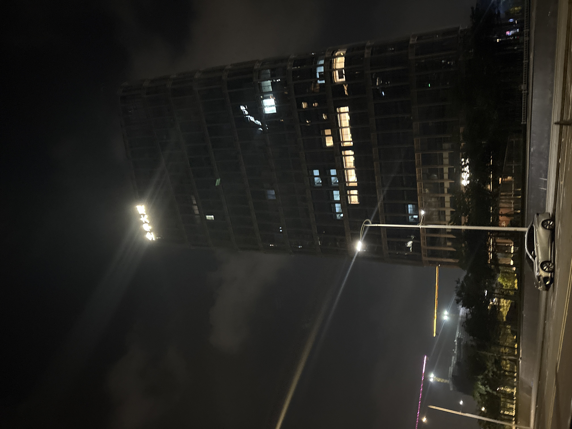 The image depicts a nighttime scene in the Haizhu District of China. A tall, modern skyscraper with a dark, glass facade dominates the center of the frame. The building is illuminated by numerous lit windows, creating a grid-like pattern of light against the dark sky. At the very top of the skyscraper, a lit sign with Chinese characters is visible. A street light with a bright beam of light cuts across the left side of the image, casting a long streak of light towards the sky and partially illuminating the building's facade. In the foreground, a silver sedan is parked on a road. Beyond the skyscraper, other buildings and construction cranes are faintly visible in the distance, with some lights from those structures also appearing in the dark sky. Trees and other vegetation are present in the middle ground, between the road and the skyscraper. The overall atmosphere is urban and nocturnal, with a focus on the architecture and the interplay of light and shadow.