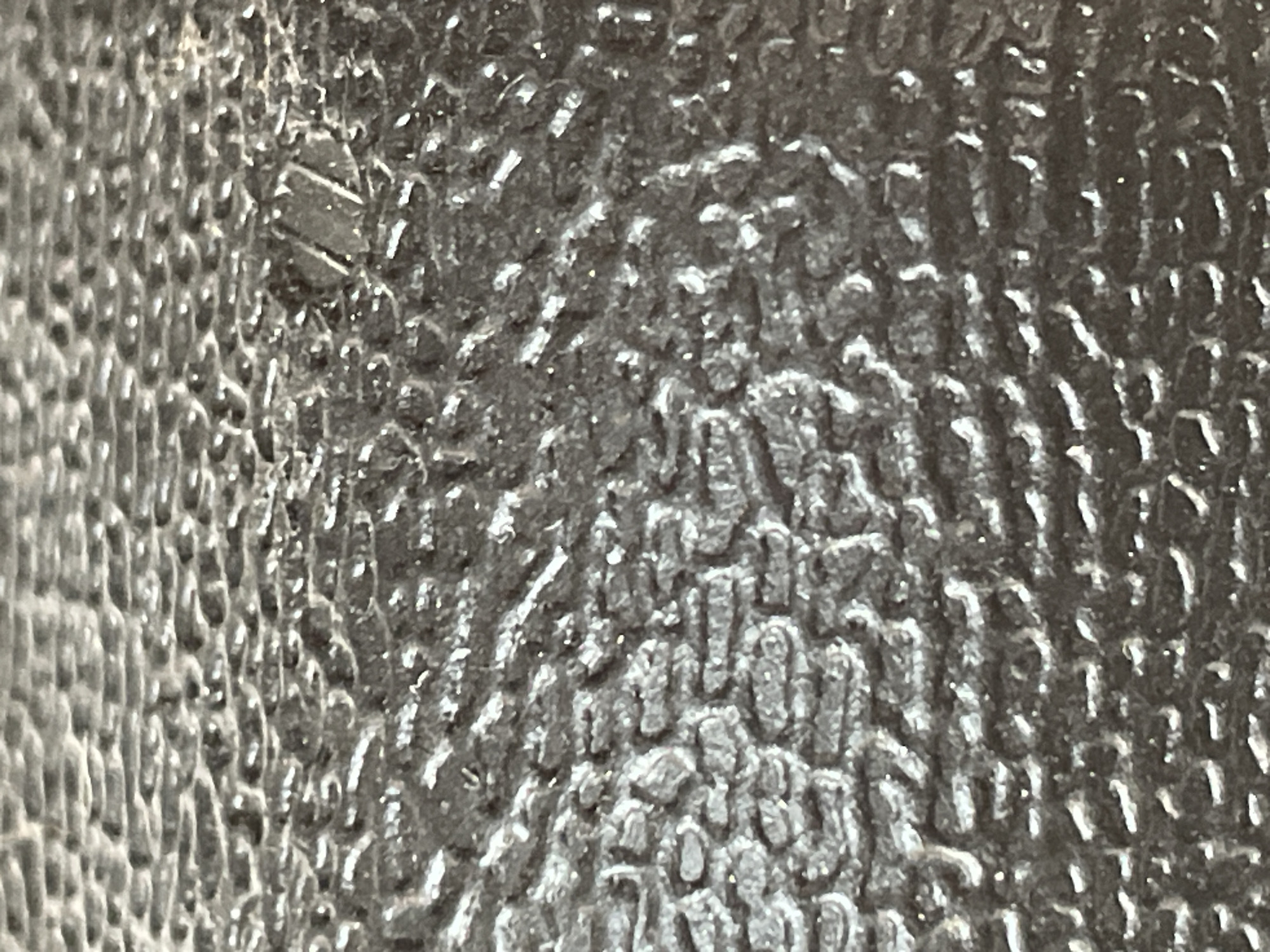 The image shows a close-up, abstract view of a textured, dark gray surface, possibly a rubberized material or a textured metal. The surface has an irregular, raised pattern, creating a play of light and shadow. There are no discernible features that would indicate a specific location within the city of Omsk, Russia, or any other urban environment. The image is purely a study of texture and light reflection on an unidentified object.
