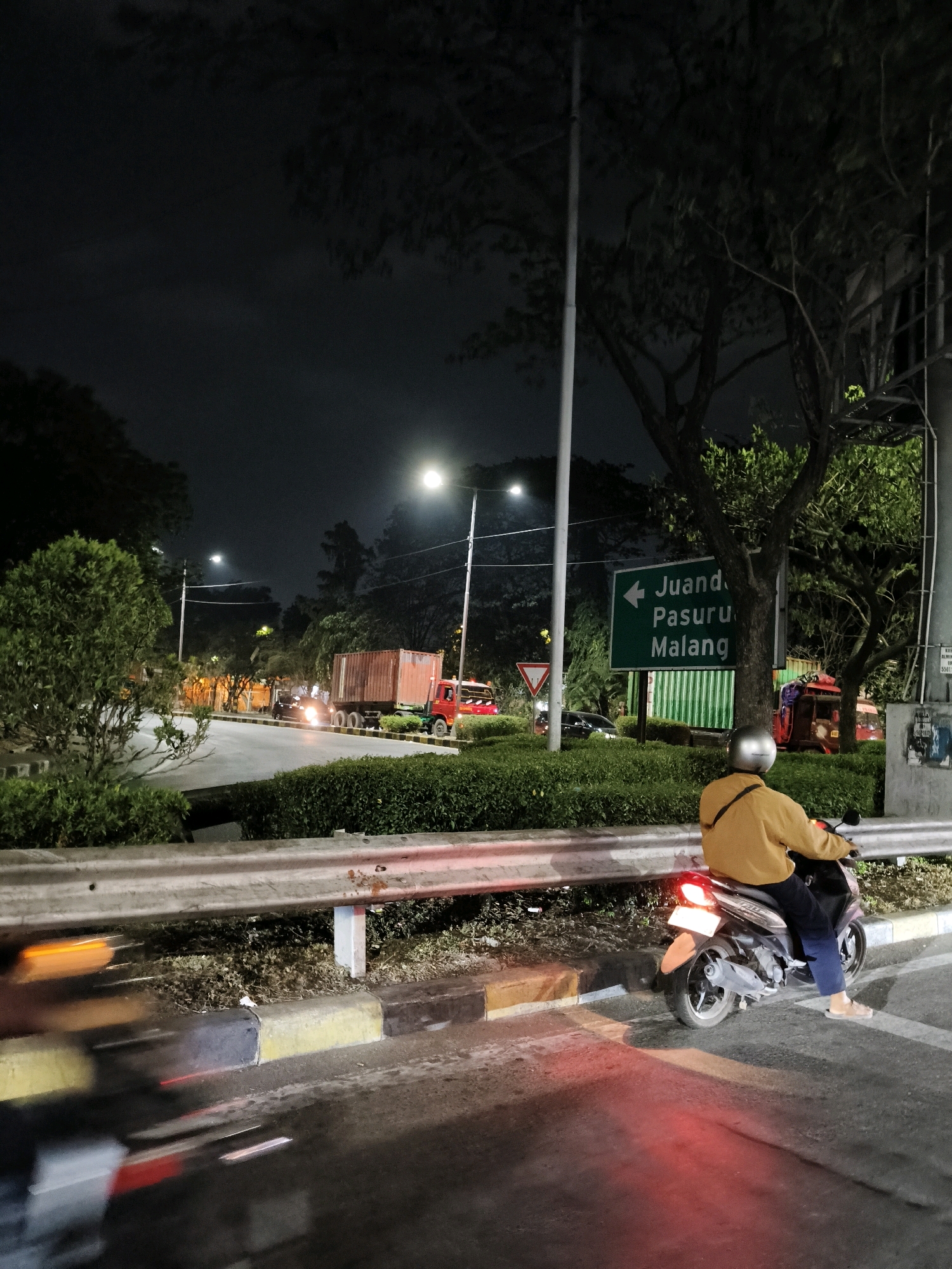The image shows a nighttime street scene in Surabaya, Indonesia, likely on a highway or major road with multiple lanes. A person is riding a scooter, wearing a yellow jacket and dark pants. The scooter's taillight is illuminated red. A road sign with green background and white text indicates directions to "Juanda," "Pasuruan," and "Malang." In the background, there are several vehicles, including a large red truck with a shipping container, a smaller red vehicle resembling a tuk-tuk, and other cars. Streetlights are visible, illuminating the road and surrounding trees. The area features a concrete barrier, a well-maintained green hedge, and a sidewalk with a yellow and black striped curb. The overall atmosphere is that of a busy urban environment at night.