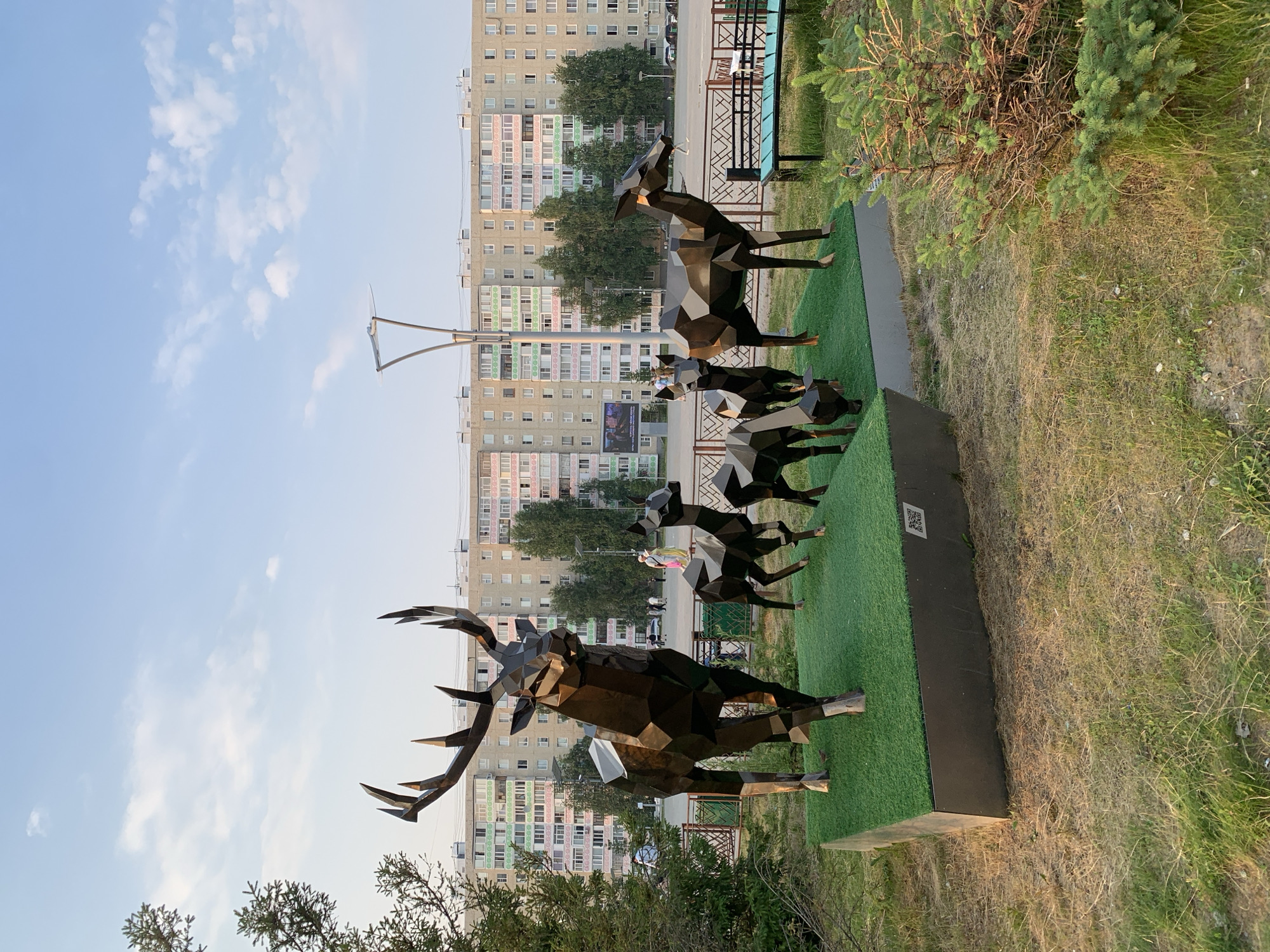 This image showcases a sculpture of a stag and a group of deer, crafted from faceted metal. The stag sculpture, prominently displayed in the foreground, is detailed with large antlers. Behind it, several smaller deer sculptures are positioned in a line, appearing to be in motion. The sculptures are set against a backdrop of a residential area, featuring tall, multi-story apartment buildings with rows of windows. Trees and greenery are visible around the sculptures and in front of the buildings. The scene is outdoors, under a bright sky with scattered clouds, suggesting daytime. A lamppost stands tall in the background. There is a QR code visible on a black base in front of the sculptures, which is an object that could provide more information about the artwork or its location. The overall impression is that of an urban art installation in a community setting.