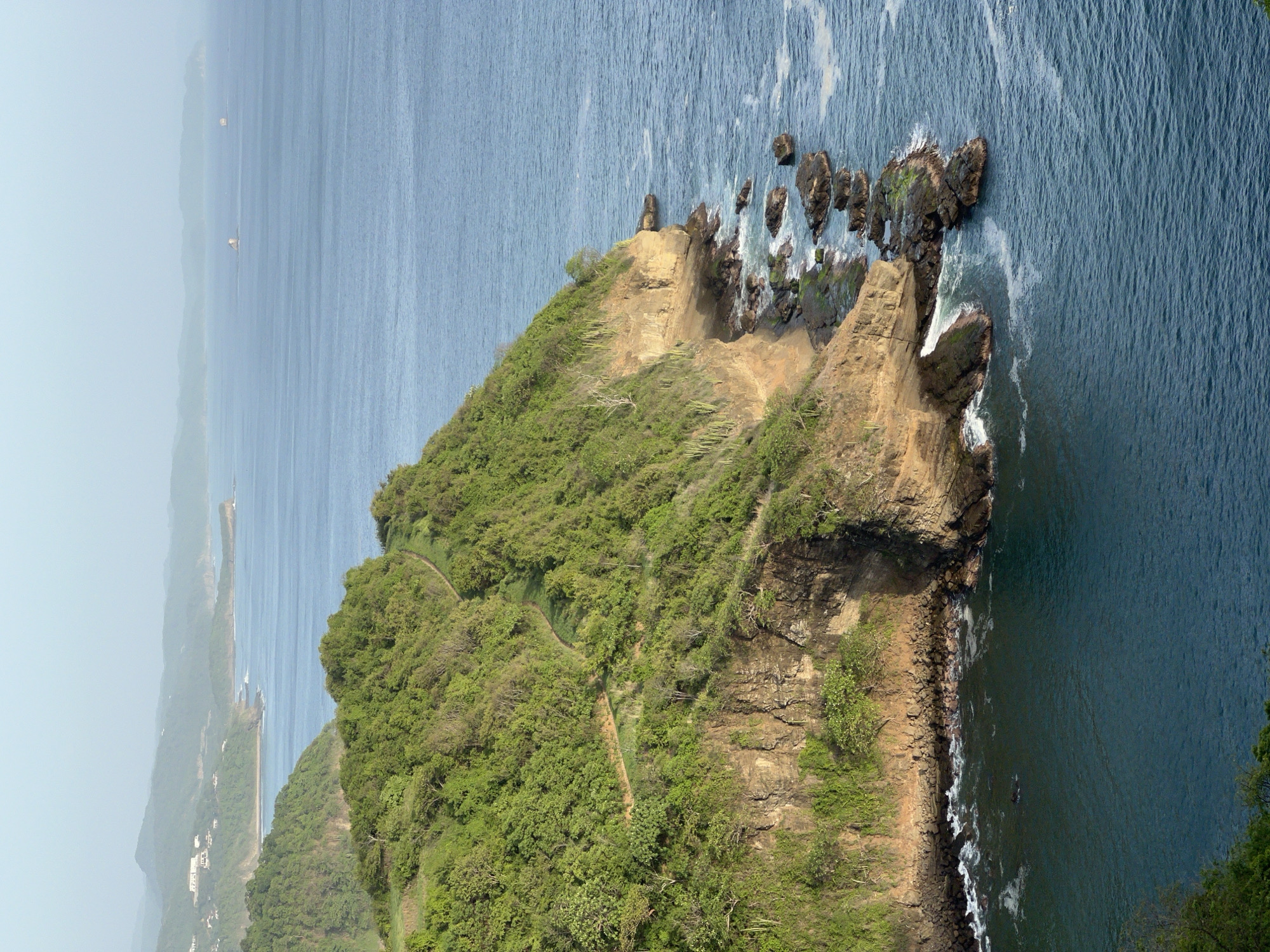 The image displays a panoramic view of the coastline near San Juan del Sur, Nicaragua. A steep, tree-covered cliff face dominates the foreground, dropping sharply into the blue ocean. Rocky outcrops are visible at the base of the cliff, with waves gently crashing against them. A winding dirt path is visible on the upper slopes of the cliff. In the background, a hazy coastline stretches into the distance, with rolling hills and a few buildings nestled on the slopes. Two small boats or buoys are visible in the far distance on the ocean. The sky is clear and light blue, suggesting fair weather.