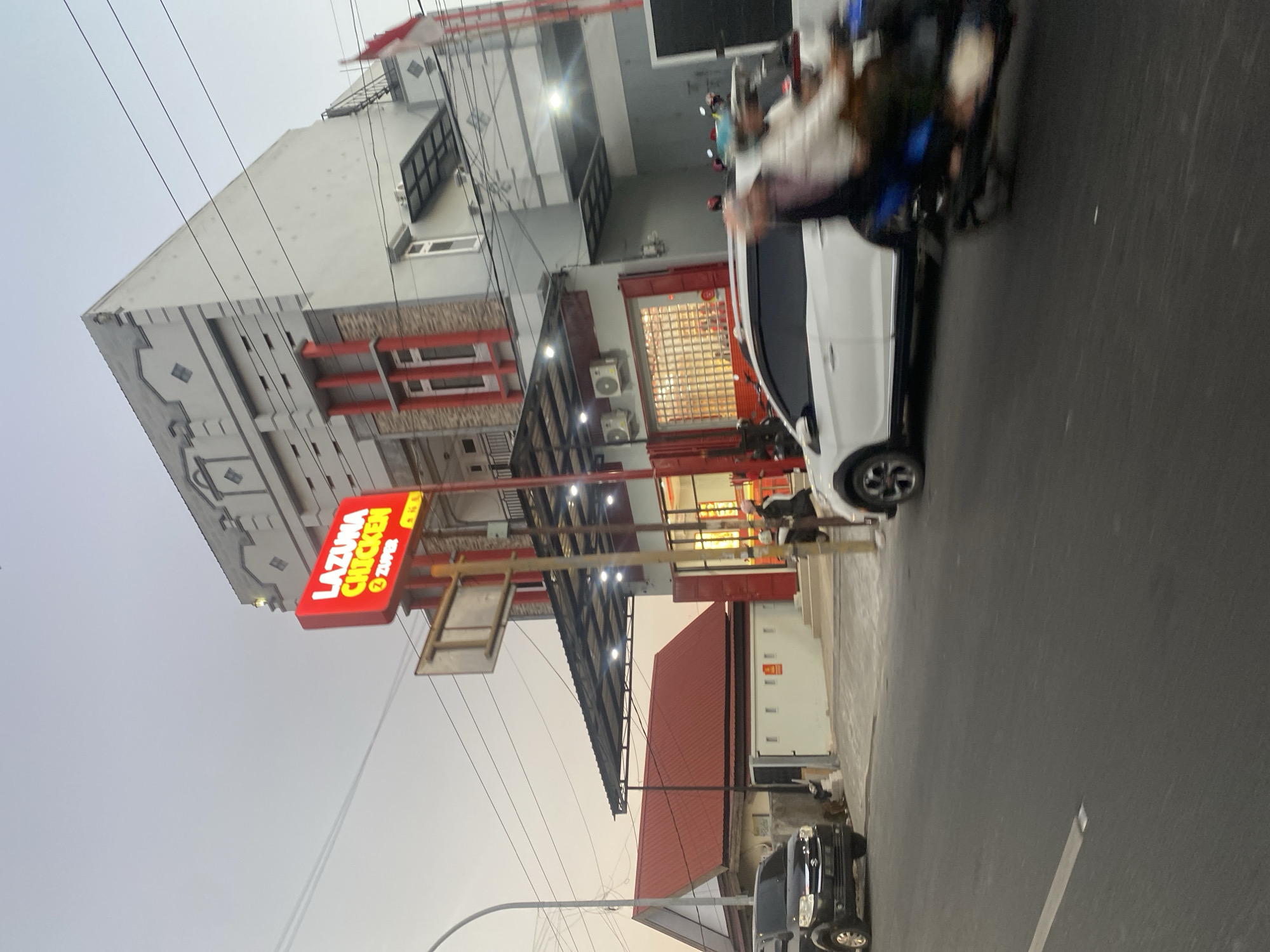The image shows a street scene in Bantaeng, Indonesia, featuring a building with a prominent "LAZUMA CHICKEN" sign illuminated in red neon. The building appears to be a commercial establishment, possibly a restaurant, with large windows and an awning. Several vehicles are visible on the street, including a black SUV parked on the left and a white car driving by, along with a scooter with two riders. The sky is overcast, suggesting it might be evening or early morning. The overall scene depicts a typical urban environment with businesses and traffic.