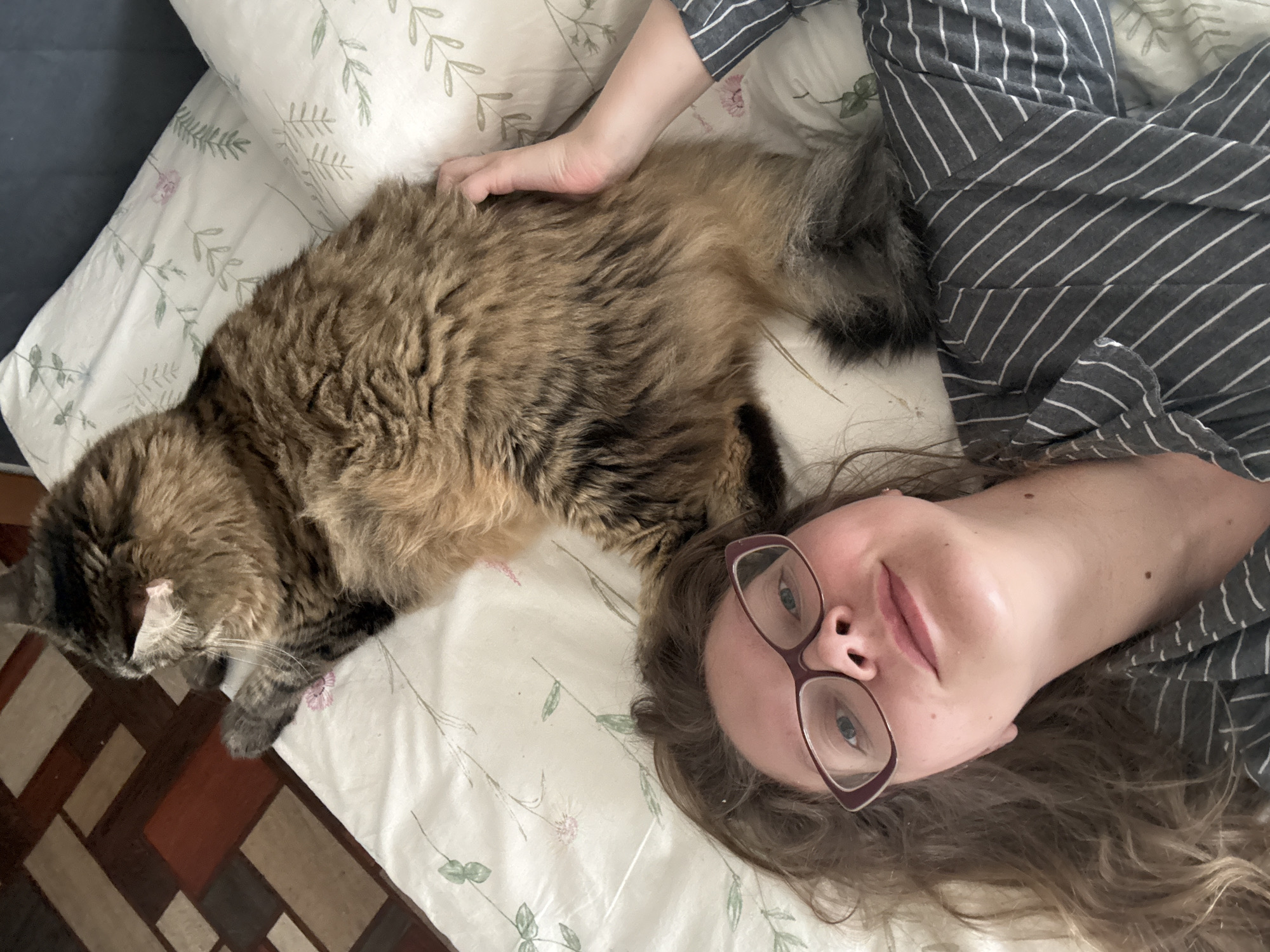 A medium shot shows a woman with brown hair and glasses lying on a bed next to a fluffy cat. The woman is looking at the camera with a slight smile. The cat is lying on its side, facing away from the camera, with its head turned to the side. The bed has white sheets with a floral pattern. The background shows a wooden floor. The image appears to be taken indoors, likely in a bedroom in Izhevsk, Russia, based on the provided context.
