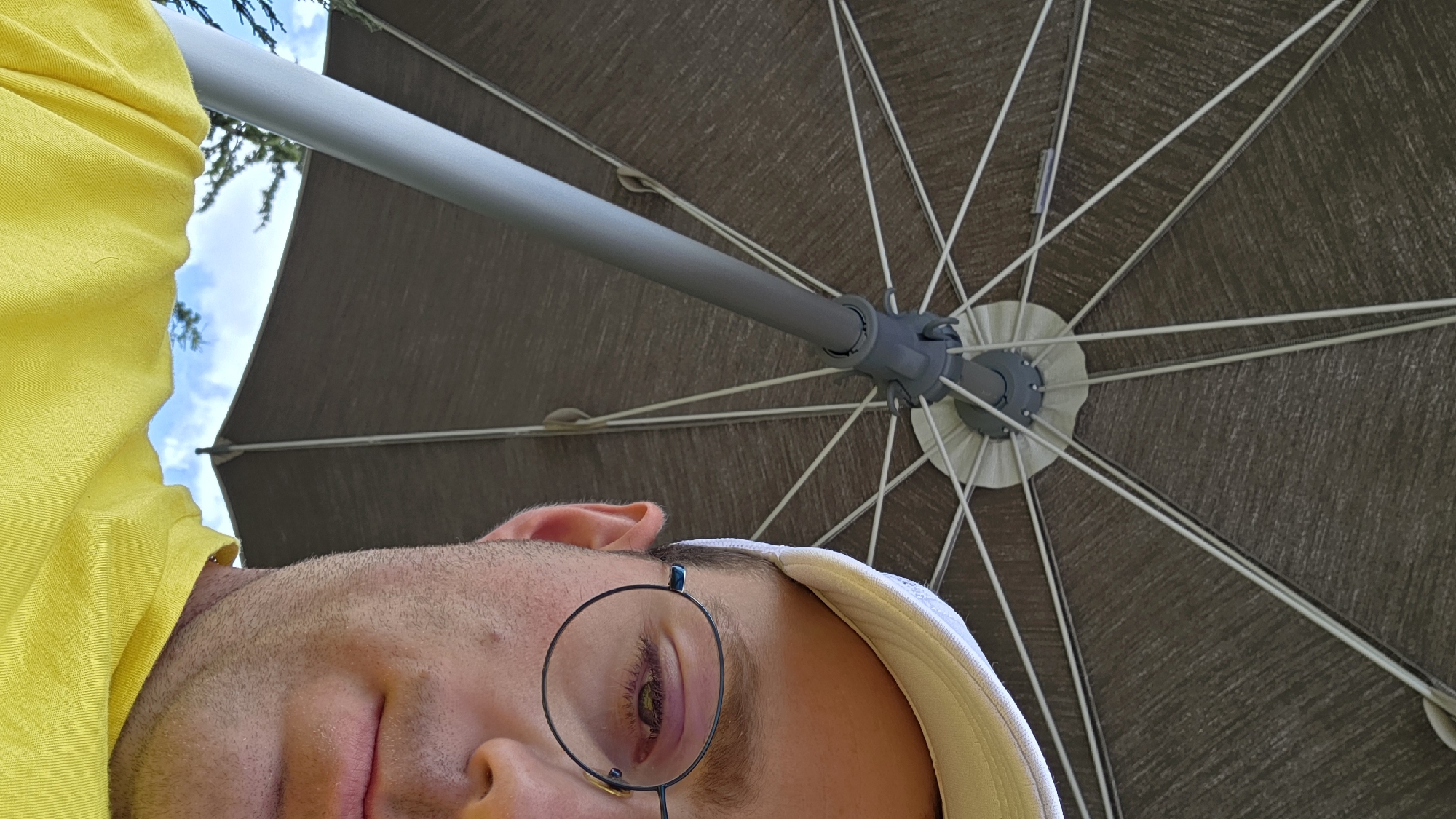 The image is an upward-facing, close-up, eye-level shot of a person wearing a white baseball cap, round glasses, and a yellow t-shirt. The person is partially obscured by an open umbrella, the underside of which is visible in the upper half of the frame. The umbrella is a dark gray color with a pattern and is supported by a light gray pole. The person's right eye is visible through their glasses, reflecting the sky and some tree branches. The sky is visible beneath the umbrella's edge, showing blue sky with some white clouds and greenery.