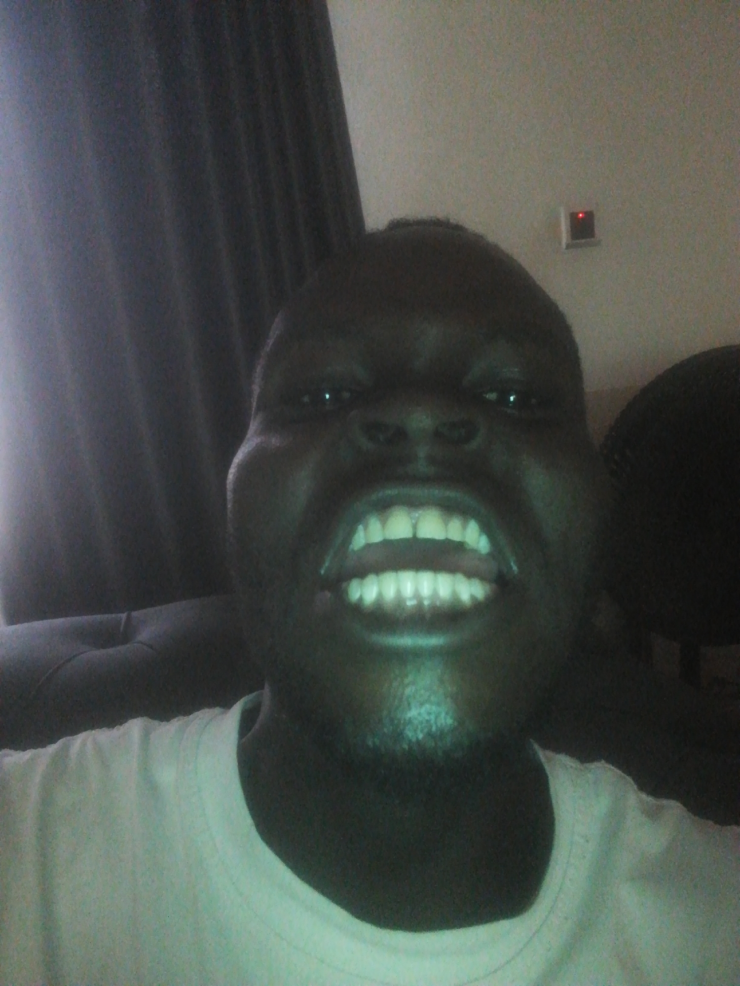 A close-up selfie of a dark-skinned person with visible teeth. The person is wearing a light-colored shirt. A dark curtain is visible in the background to the left. An electronic device with a red light is on the wall in the background. Part of a fan is visible on the right. The location is possibly Ikorodu, Nigeria.