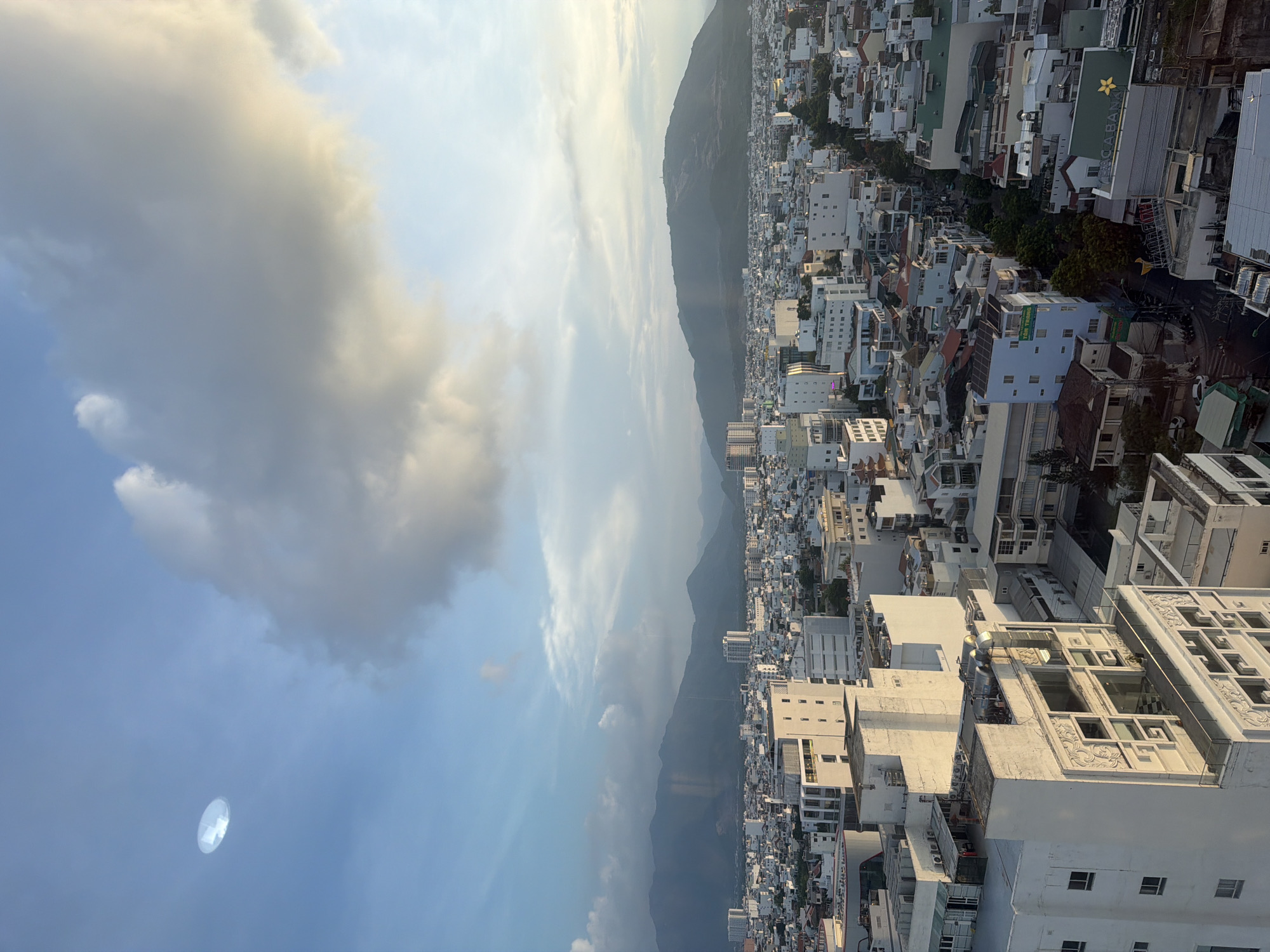 The image depicts a panoramic view of a city, likely Nha Trang Ward, Vietnam, from an elevated perspective. The foreground is dominated by a dense collection of white buildings, varying in height and architectural style, suggesting a mix of residential and commercial structures. Some buildings feature modern designs with balconies and large windows, while others appear older and more traditional. Rooftops are visible, some adorned with air conditioning units or solar panels.
In the mid-ground, the city continues to sprawl, with smaller buildings and glimpses of streets and vegetation. The urban landscape is backed by a range of undulating, verdant mountains that fade into a hazy, blue sky.
The sky itself is a prominent feature, with a mix of bright blue sky and soft, diffused sunlight filtering through scattered clouds. A large, oval-shaped, translucent object is visible in the upper left quadrant of the sky, reflecting light and appearing to be suspended in the air. The overall lighting suggests either early morning or late afternoon, with warm tones illuminating parts of the clouds and mountain ranges.
The scene is devoid of visible human activity or any direct interaction between subjects or objects, presenting a static, aerial snapshot of the city and its natural surroundings under a dynamic sky.