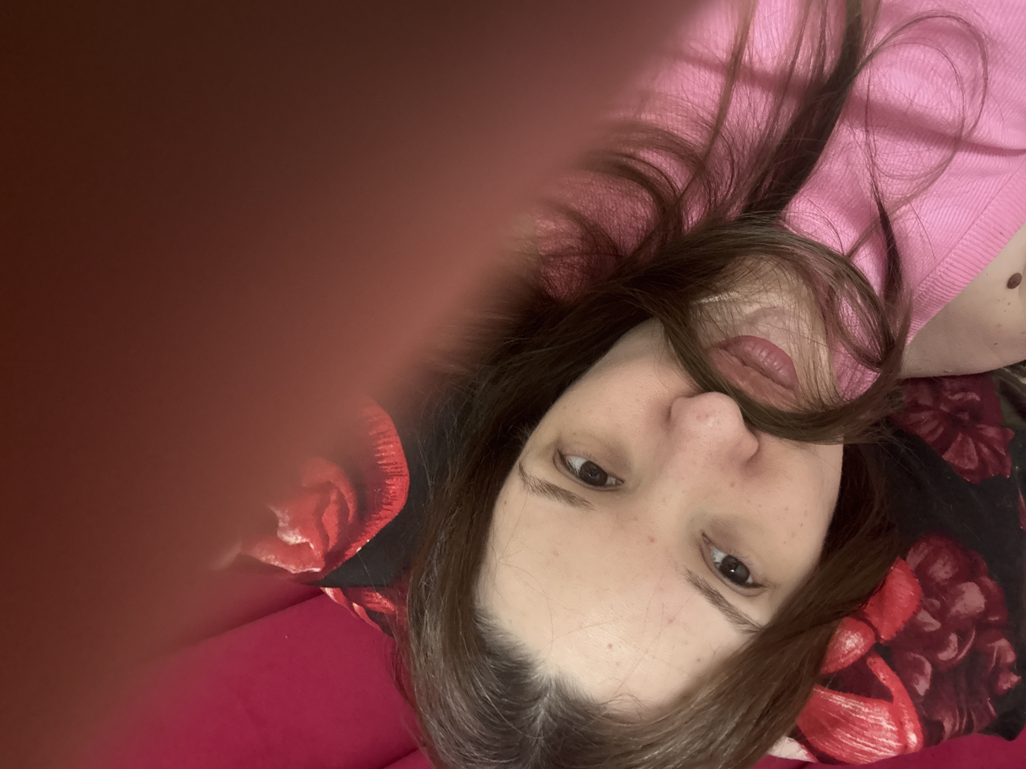 This is a close-up, low-angle, overhead shot of a young woman lying down, looking upwards at the camera. She has long, dark brown hair that is partially obscuring her face and is spread around her head. She is wearing a pink ribbed knit top. Her skin is fair with a scattering of freckles on her cheeks and nose. Her lips are parted slightly, and she has brown eyes. She is lying on a surface that appears to be a bed or couch covered with a red floral patterned fabric and a pink blanket or sheet. The background is blurred, with hints of red from the floral pattern and pink from the fabric. The lighting is soft and warm, suggesting an indoor setting. There are no visible text elements in the image. The overall mood is casual and intimate. The location context of Засечное, Russia is not visually discernible from the image itself.