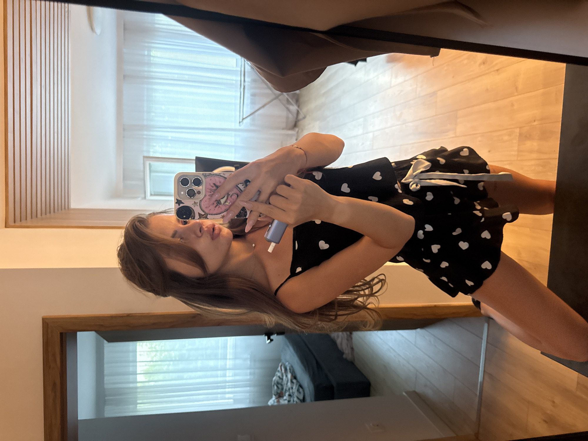 A young woman with long, brown hair is taking a mirror selfie in what appears to be a bedroom or dressing area. She is wearing a black spaghetti strap top adorned with white hearts and white drawstring detailing at the waist. The woman is holding a smartphone in her right hand, which has a pink case with various decorative stickers, including stars and celestial motifs. Her left hand is raised, holding a small, light purple cosmetic product, possibly a serum or applicator, near her face.
The setting is a room with wooden floors and walls. A large mirror reflects the woman and the environment behind her. In the reflection, we can see a window with white blinds, suggesting it is daytime. A dark couch with a patterned throw blanket is visible in the background reflection, indicating a living space or a bedroom. The lighting suggests it is likely daytime. The woman's expression is neutral, focused on taking the photo. The overall impression is casual and personal, a typical mirror selfie taken in a domestic setting. The location is Plovdiv, Bulgaria.