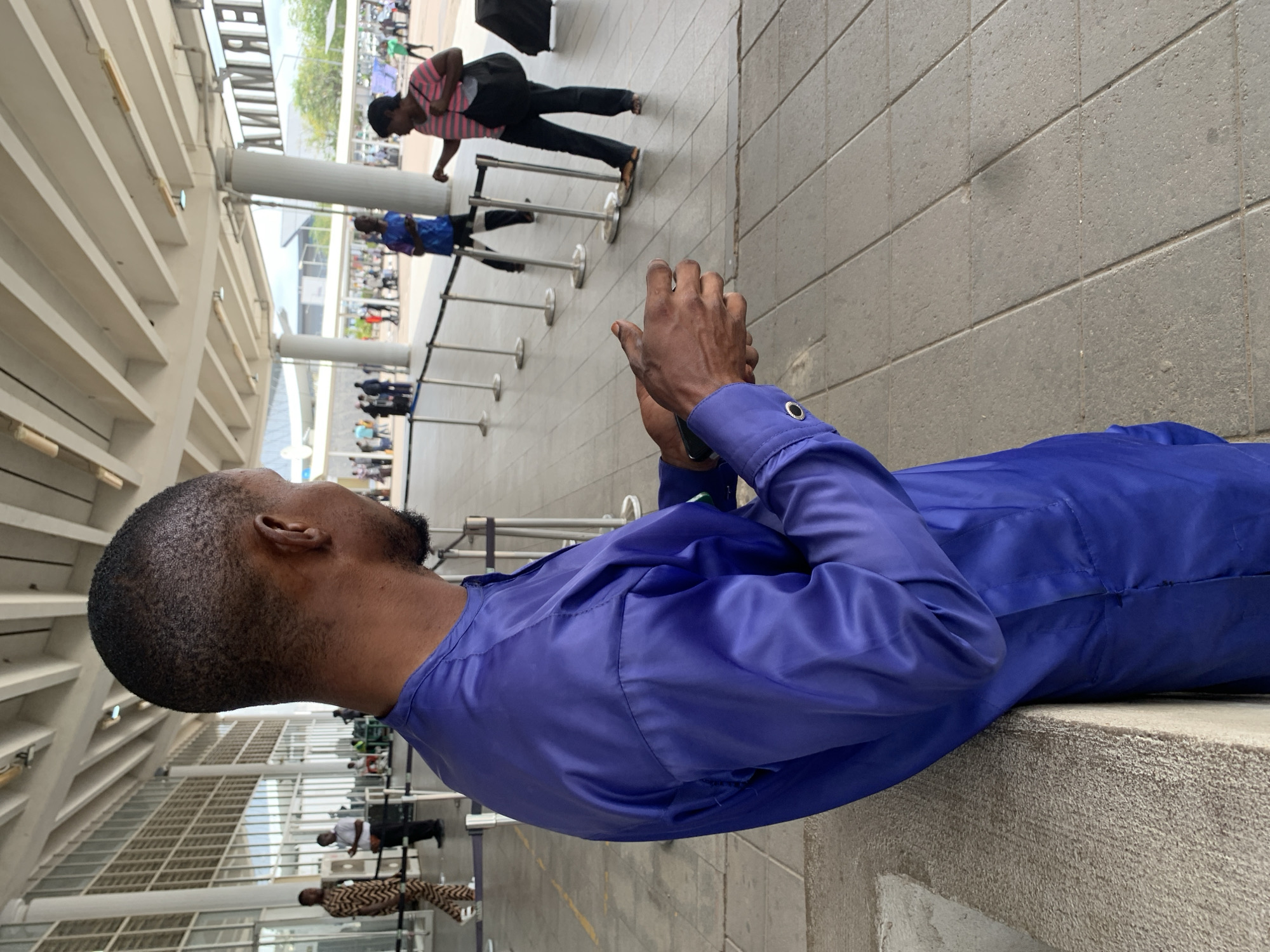 The image captures a scene at what appears to be a busy airport terminal or a large public transportation hub in Abuja, Nigeria, during daytime.
In the immediate foreground, a man is seen from behind and slightly to his right, resting his left arm on a low, textured concrete ledge. He has short, dark hair and a beard, and is wearing a long-sleeved, royal blue or purple satin-like shirt, possibly a traditional Nigerian garment. His right hand is holding a mobile phone or similar small device, indicating he is occupied with it while waiting.
The background reveals a spacious and bright architectural environment with high ceilings supported by large concrete pillars and an intricate structure of horizontal beams or girders. This design suggests a modern, open facility, likely allowing natural light to filter in. In the mid-ground, a queue is forming, demarcated by a line of silver stanchions. A woman wearing a pink and white striped top, black trousers, and a black backpack is visible in this queue, with a black rolling suitcase placed near her. Another person in a blue shirt is further down the line. Numerous other individuals can be seen in the distant background, contributing to the sense of a busy public space.
Prominently displayed on a building facade in the upper background, the word "TERMINAL" is visible, horizontally oriented but inverted, confirming the location as a transport terminal. The overall atmosphere depicts people engaged in typical travel-related activities, likely waiting for check-in, security, or boarding. A small tree is also visible in the distant background, suggesting an outdoor or semi-outdoor section of the facility.