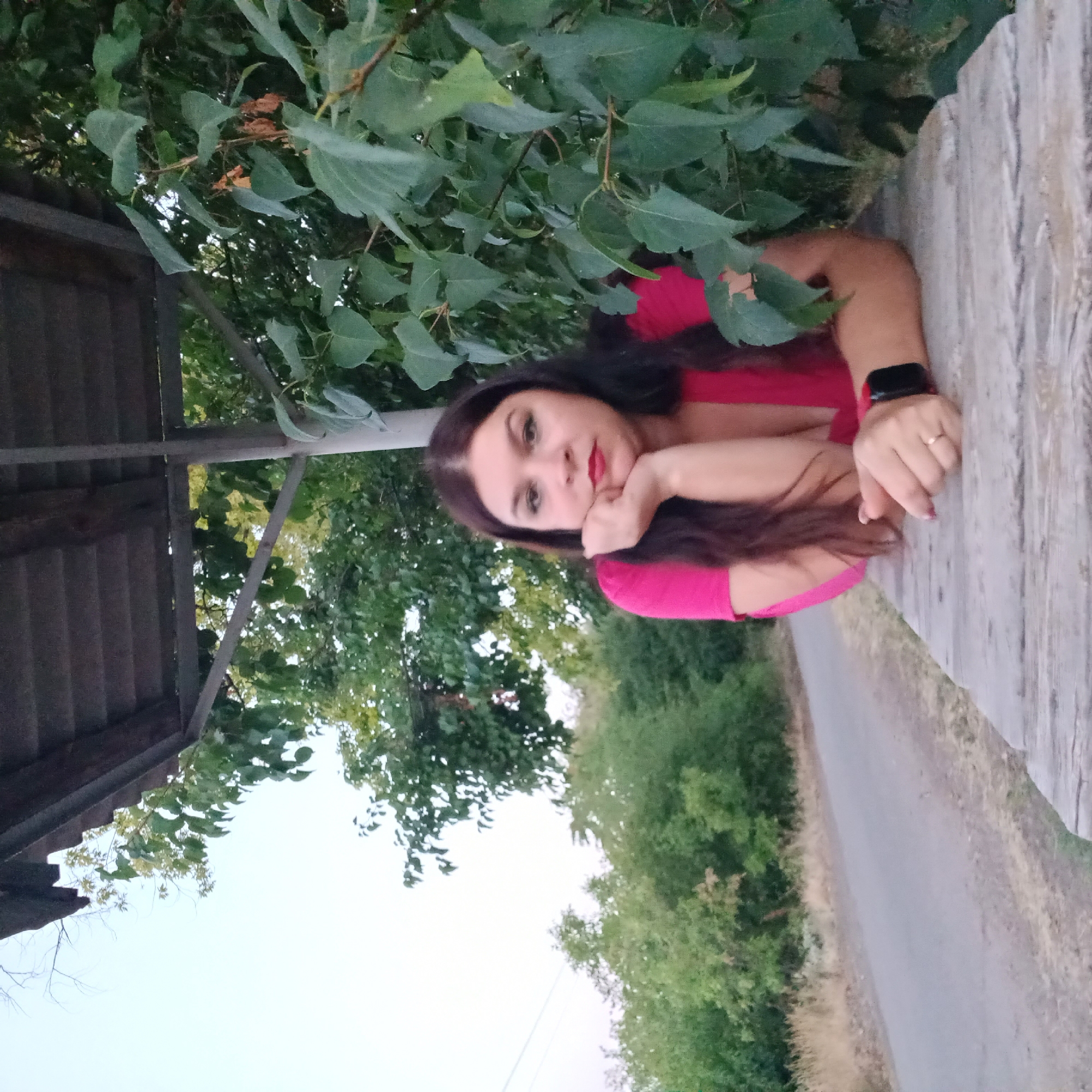 In Berezhynka, Ukraine, a woman with dark hair and red lipstick leans on a wooden surface under a covered structure. She wears a bright pink top. Her right hand is propped against her chin, and her left arm rests on the surface. A smartwatch is visible on her left wrist. The background consists of lush green trees and a light-colored sky. A paved road is visible to the left, bordered by dry grass. The scene appears to be outdoors, possibly at a rest stop or picnic area.