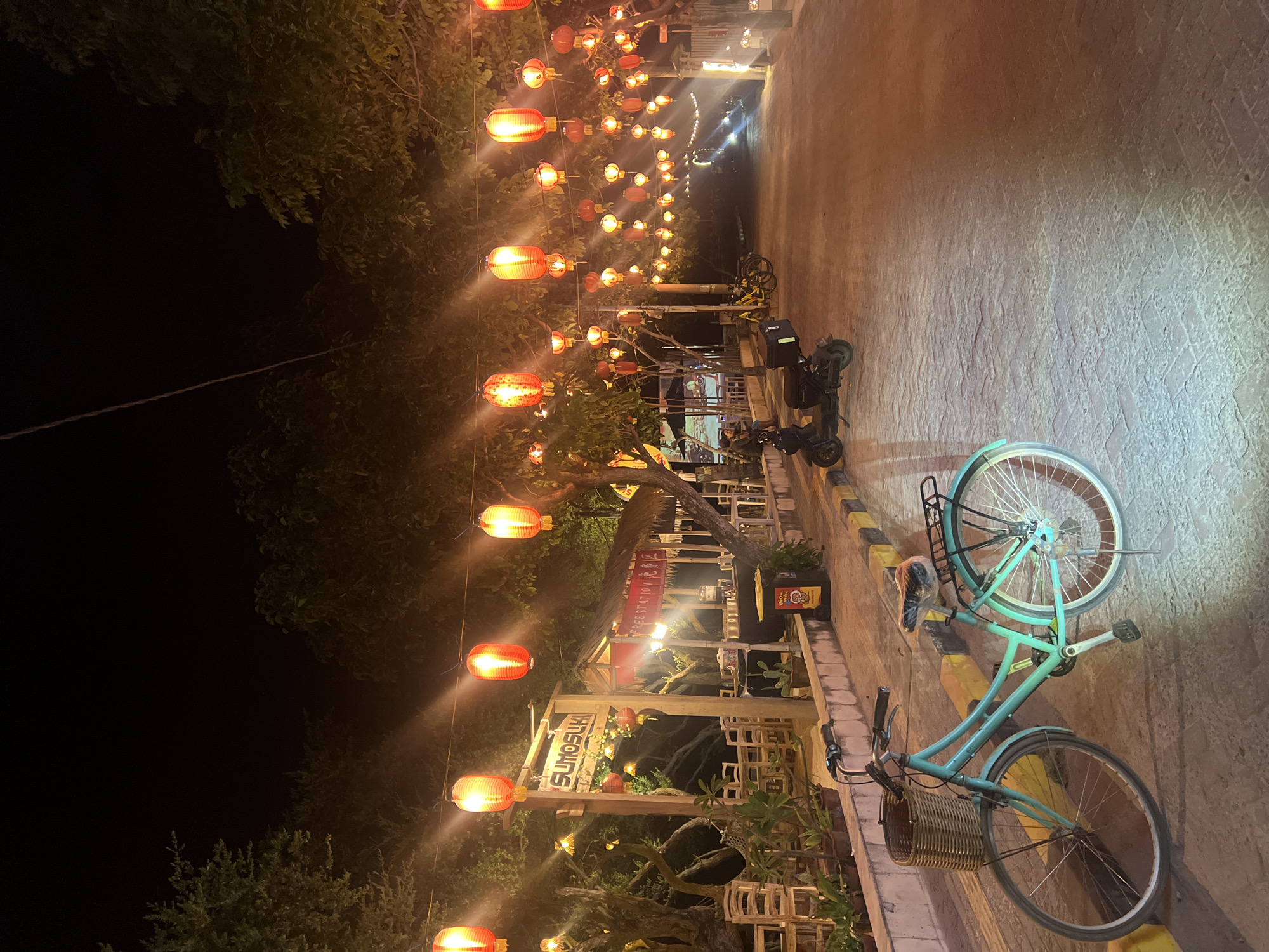 The image captures a vibrant night scene along a street or pathway, illuminated primarily by numerous red and orange paper lanterns strung overhead. The time of day is clearly night, with a dark sky visible between the illuminated foliage of trees lining the left side of the path.
In the foreground, a turquoise bicycle with a wicker basket attached to the handlebars and a rack over the rear wheel is prominently parked on the right side of the path. Its light-colored frame stands out against the dark ground. Further down the path, several scooters or motorbikes are also parked. The path itself appears to be paved with light-colored bricks or cobblestones, showing texture and shadows cast by the various light sources.
On the left side of the path, beneath the canopy of trees and lanterns, there are structures suggestive of outdoor eateries or stalls. A prominent wooden sign is visible, displaying the text "SUMOGLUKL" in a stylized font. Further along, there's another structure with visible text that appears to say "COFFEE STATION". Outdoor seating arrangements, including chairs and tables, are scattered around these establishments. A yellow bucket with "Wonderfull" and some red text (possibly "HOT") is also seen near one of the stalls.
The overall atmosphere is inviting and festive, created by the warm glow of the lanterns. The setting appears to be a lively commercial area or a decorative street, possibly in Southeast Asia, given the style of lanterns and the presence of scooters.