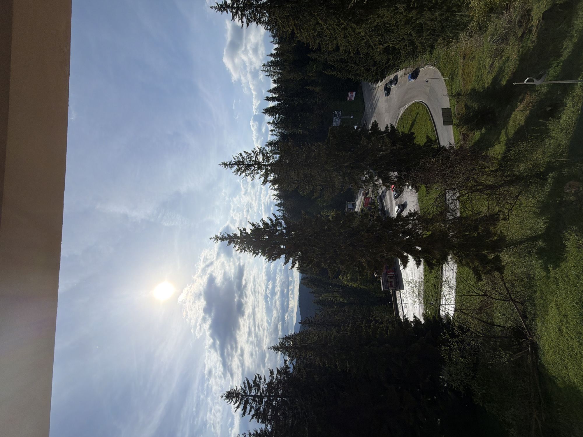 A ground-level, medium shot shows a winding asphalt road with several vehicles traveling in both directions. The road is bordered by evergreen trees, grass and some buildings. The sky is partly cloudy with the sun visible. A building is located to the left, and the location is near Progled, Bulgaria.