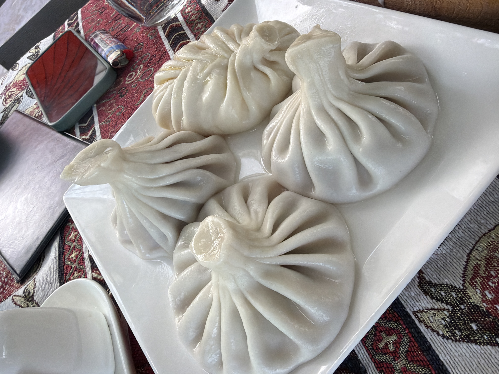 This is a close-up, overhead shot of a white ceramic plate containing four khinkali, a traditional Georgian dumpling, served on a table with a red and white patterned tablecloth. The khinkali are plump and appear to be freshly steamed, with a visible broth pooling at the bottom of each one. The dough is a pale, almost translucent white, and the pleated tops are gathered to a point. To the left of the plate, a white ceramic teacup and saucer are partially visible. In the background, a dark, rectangular object, possibly a menu or a tablet, rests on the tablecloth, next to a red object that resembles a lighter. The lighting is bright, suggesting it's daytime, and the overall scene is indoors. The image was taken in Kaluđerac, Montenegro.