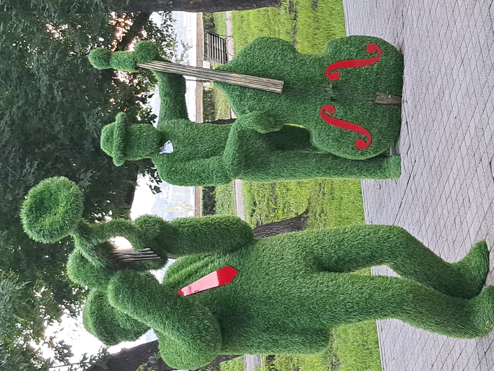 This image depicts two grass-covered sculptures of musicians in a park setting. The sculpture on the left, dressed in a red tie, appears to be playing a wind instrument and possibly wind chimes. The sculpture on the right, wearing a hat, is positioned behind a grass-covered double bass with red decorative f-holes. Both figures are abstract and anthropomorphic, with rounded, fuzzy textures. The background shows trees, grass, a paved walkway, and a bench. The lighting suggests it is daytime, possibly late afternoon due to some dappled sunlight. The image is taken from a medium distance, capturing both sculptures and their immediate surroundings. There is no visual information within the image to suggest the location is Irkutsk, Russia.
