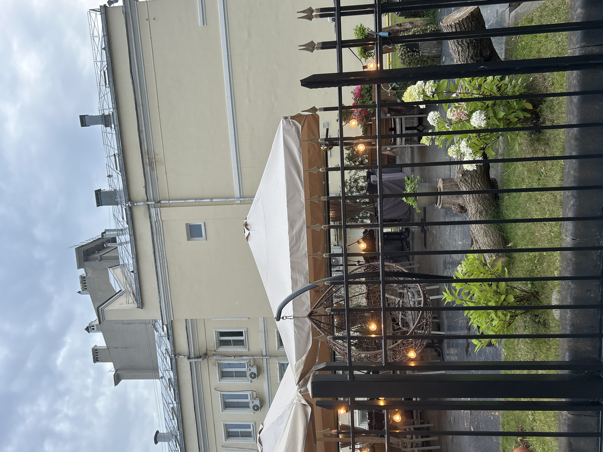 A cloudy, overcast day in an outdoor dining area of an unknown city. The foreground features a black metal fence with vertical bars. Beyond the fence, a large, woven hanging chair is suspended from a curved metal pole. The chair is empty. Several beige umbrellas are set up, providing shade over tables and chairs. String lights with Edison-style bulbs are draped around the area and also hang from the umbrellas, casting a warm glow. In the background, a light-colored building with multiple windows and air conditioning units is visible. The building has a flat roof with several chimneys and what appear to be ventilation shafts. The ground is paved, and there are some potted plants and decorative logs visible in the foreground on a grassy area.