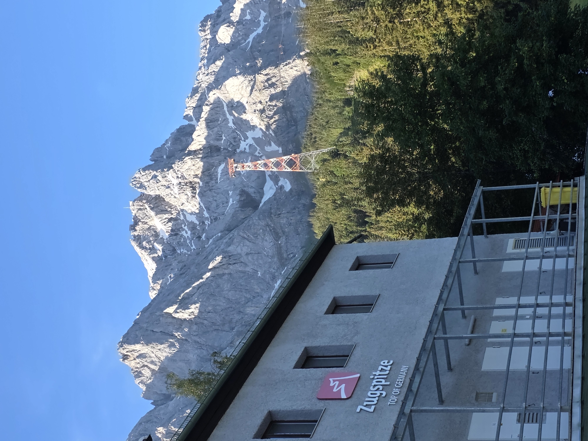 The original image, taken from an outdoor perspective, features a building in the foreground and a majestic mountain range in the background. The building, with its light-colored facade, displays the "Zugspitze TOP OF GERMANY" logo prominently. It has multiple windows and an exterior metal walkway with railings and doors. To the right of the building, a dense forest of evergreen trees stretches towards the mountains. A tall, red and white tower, characteristic of a ski lift or cable car support, stands amidst the trees, with cables extending upwards towards the mountain peaks. The mountains themselves are rugged and imposing, with visible snow patches on their upper slopes, indicating a high altitude. The sky above is a clear, vibrant blue. The scene is captured in daylight, with sunlight illuminating the mountain tops and casting shadows, suggesting it's either early morning or late afternoon.