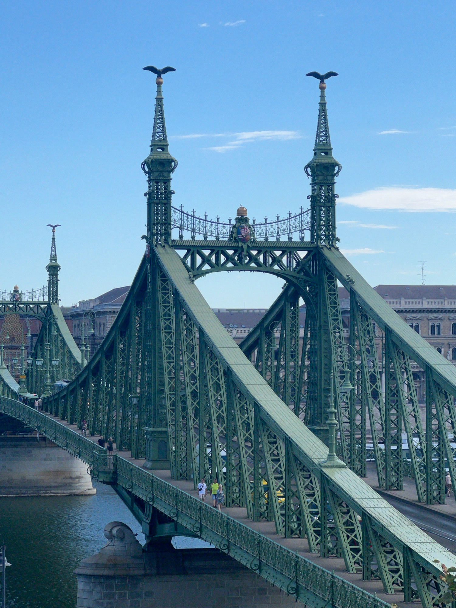 The image displays the Szabadság híd, or Liberty Bridge, in Budapest, Hungary, a distinctive green suspension bridge spanning the Danube River. The bridge's intricate metalwork is prominent, with its large, latticed trusses and ornate lamp posts. Two prominent eagle statues stand atop tall spires at either end of the bridge's central arch, their wings spread wide. In the background, traditional European architecture with tiled roofs and arched windows lines the riverbank. Several pedestrians stroll across the bridge, some of whom are walking or cycling. The water of the Danube is visible below, reflecting the blue sky and white clouds. The lighting suggests it is daytime, with the sun illuminating the scene.