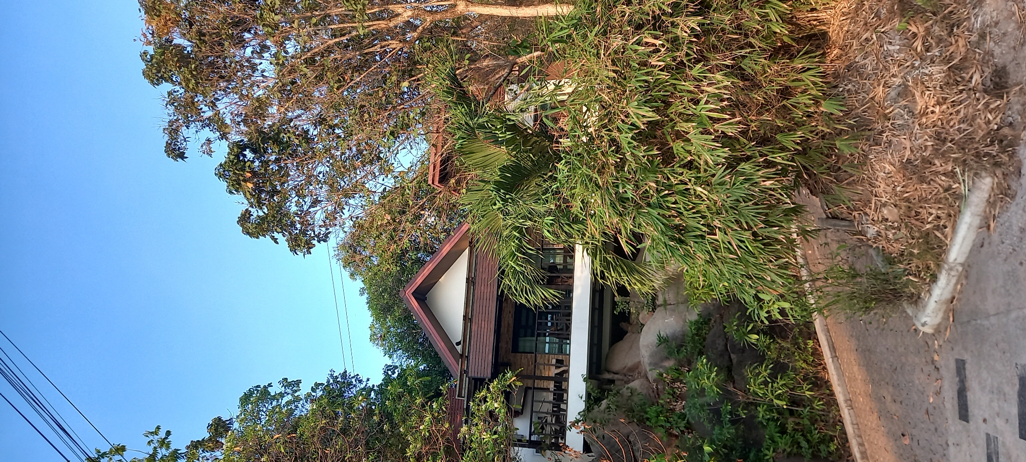 The image shows a building with a red-trimmed gable roof in Baan Wok Tum, Thailand. The building is surrounded by trees, including bamboo, and is situated on a rocky outcrop. The sky is clear blue. A concrete road is visible in the foreground. Power lines are visible in the top left.