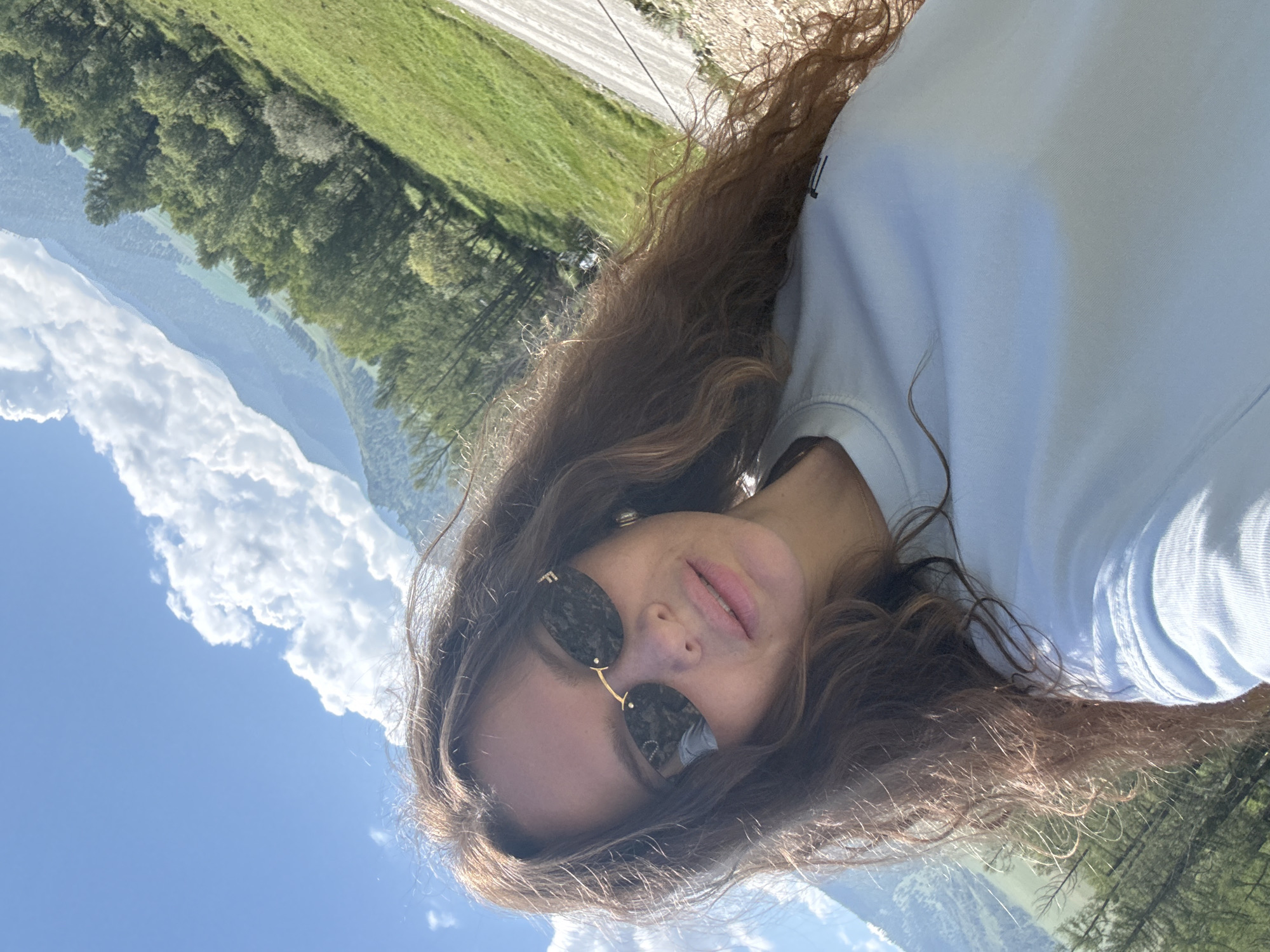 A medium shot shows a young woman with long, wavy brown hair, wearing dark sunglasses and a light blue t-shirt, posing for a selfie outdoors. She is looking at the camera with a slight smile. Her hair is partially covering her face and shoulders. Behind her, a vast landscape unfolds under a clear blue sky with fluffy white clouds. Rolling green hills covered with trees dominate the background, and mountains are visible in the distance. The terrain appears to be natural and undeveloped, suggesting a rural or mountainous setting. The image does not provide specific details that can identify the location as Bichiktu-Boom, Russia.