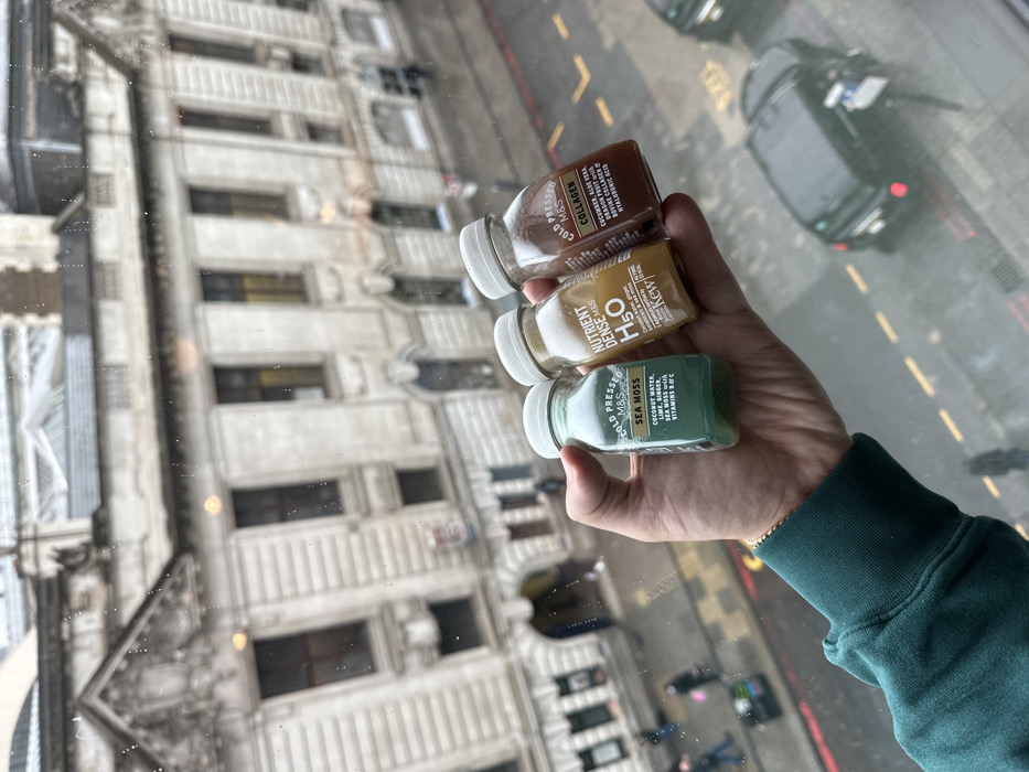 The image depicts an elevated view through a rain-splattered window, overlooking a street and a historical building in London, United Kingdom. In the foreground, a person's left hand, adorned with a gold-colored wristband and wearing a dark green sleeve, holds three small, clear plastic bottles vertically. From left to right, the bottles are labeled "COLD PRESSED M&S SEA MOSS" (green liquid), "NUTRIENT DENSE M&S H2O" (yellow/orange liquid, with a Kew Gardens logo), and "COLD PRESSED M&S COLLAGEN" (brownish-red liquid).
The background, visible beyond the wet glass, features a large, ornate, light-colored stone building with multiple windows and classical architectural elements, including pilasters and decorative pediments. An arched entrance is visible at street level on the left side of the building. The street below appears wet, suggesting recent precipitation. Several indistinct pedestrians are visible on the pavement. At least two dark-colored vehicles are present on the street; one is a dark SUV-type car. The sky is overcast and grey.
