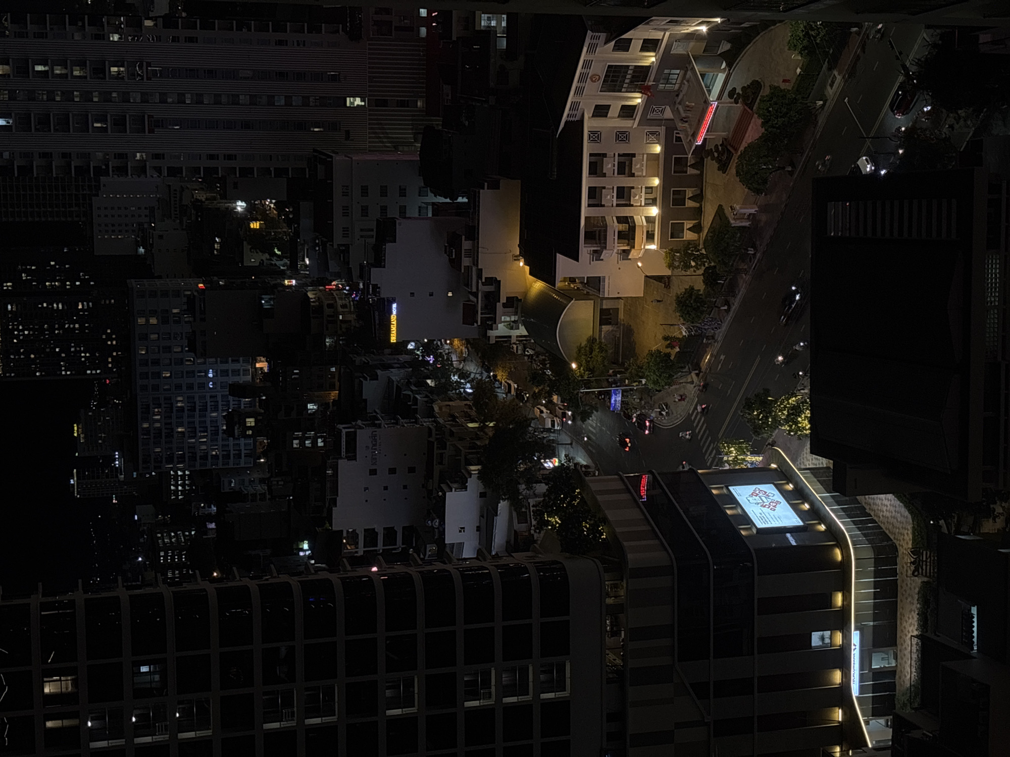 This is an aerial, night-time, medium-shot photograph of a street scene in Nha Trang Ward, Vietnam. The composition is dominated by the dark silhouettes of buildings and the illuminated street below, punctuated by the lights of vehicles and buildings.
In the foreground, on the left, is a large, dark building with a curved facade, its windows appearing as dark rectangles. Below this, a more modern building with a horizontal display screen is visible. The screen shows abstract graphics and text, with a prominent logo that appears to be for "TPBANK". The lower section of this building has a horizontal illuminated sign that reads "TPBANK" with a small emblem.
To the right of the foreground building, a street curves away from the viewer. Cars and motorcycles are visible on the street, their headlights and taillights creating streaks of light. Pedestrians can be seen on the sidewalks. Several trees with illuminated foliage are scattered along the street.
Further down the street, a white building with multiple floors is illuminated, featuring a patterned facade and balconies. A red illuminated sign is visible at the entrance to this building. Across the street, another building is illuminated, with several windows showing interior lights.
In the background, numerous tall buildings rise into the night sky, their windows lit in various patterns, creating a sense of urban density. The overall impression is of a bustling city at night, with a focus on the interplay of artificial light and the darkness of the night sky. The scene appears to capture a typical urban environment with considerable architectural development and ongoing activity.