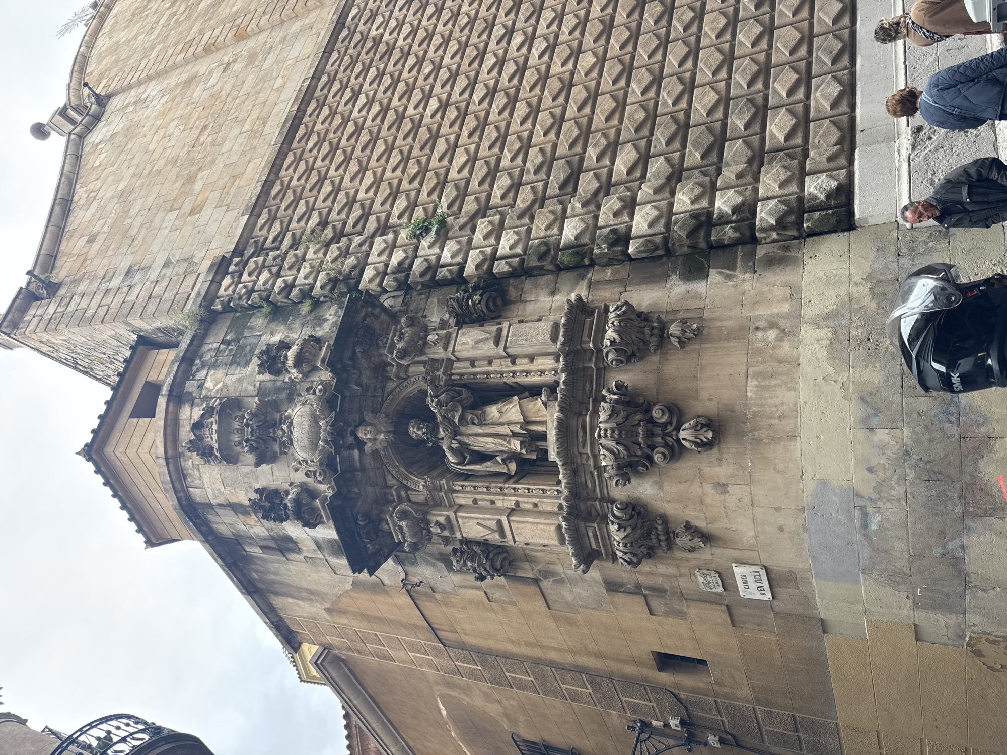 The image captures a street view in Barcelona, Spain, showcasing a section of a historical building. The building's architecture is notably characterized by its textured façade, featuring rows of diamond-shaped stones. A niche on the building houses a statue adorned with intricate decorative elements. Two small street signs, "Carrer d'en Xucla" are affixed to the building. A motorcycle helmet rests on the wall. Three pedestrians are visible near the building.