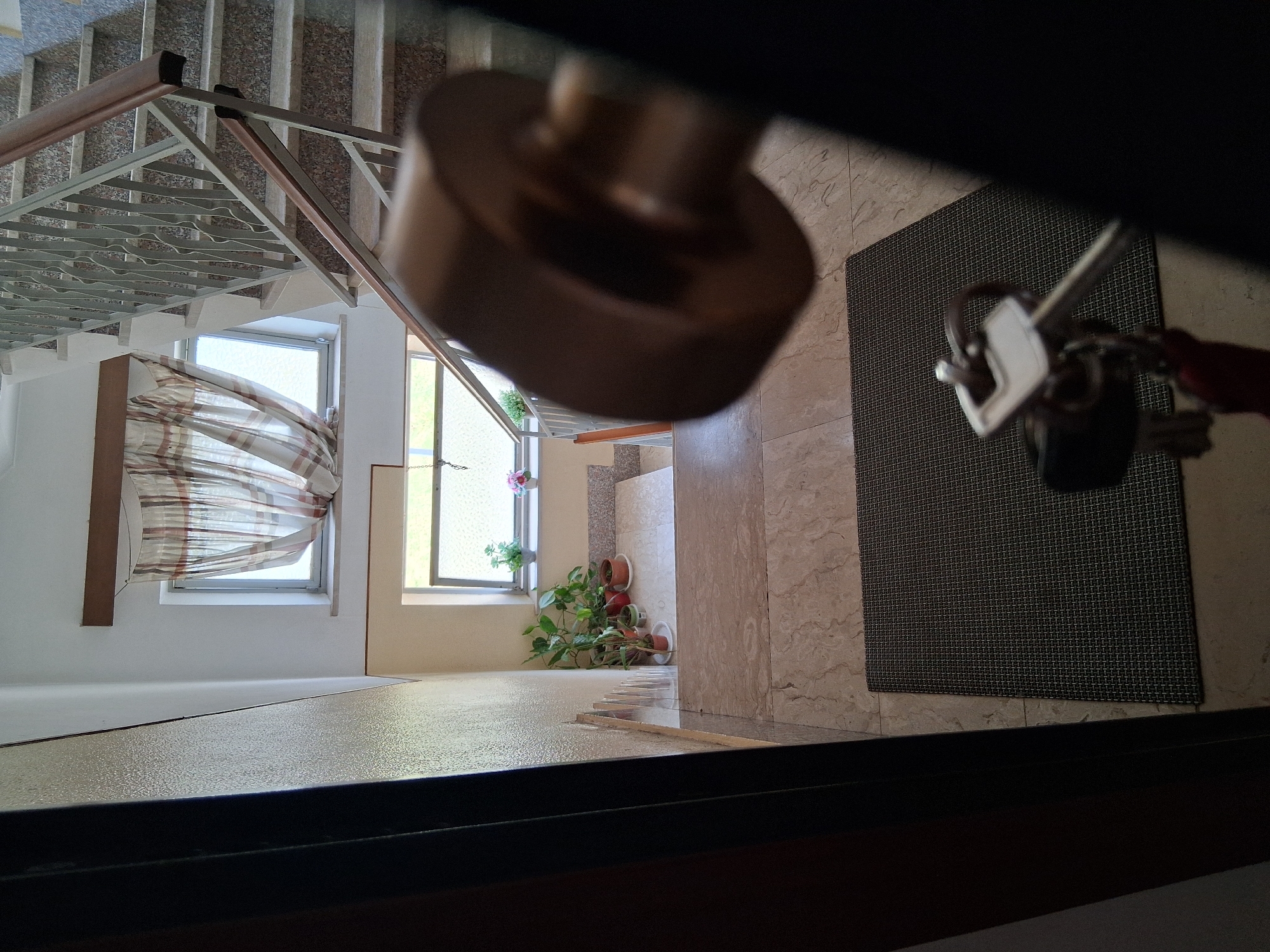 The image depicts a view from an open doorway looking into a building's interior, likely a residential building. In the foreground, a dark door frame is visible on the left, and on the right, a portion of a door handle, out of focus, occupies the lower right quadrant. A doormat, dark grey with a grid pattern, lies on the floor directly ahead. On the doormat, a set of keys with a white key fob and a red tag are visible. Beyond the doormat, the floor transitions to a lighter tiled surface.
Further into the image, a staircase with grey speckled steps and a metal railing with wooden handrails ascends. The staircase is flanked by walls. Below the first flight of stairs, a small landing area features several potted plants on a beige surface. Two windows are visible; one larger window above the plants shows a striped curtain, and a smaller window is visible below it, also with a curtain. The lighting suggests it is daytime. The location is identified as Colorno, Italy, implying this is a building within that city. The scene appears to be an interior entrance or hallway.