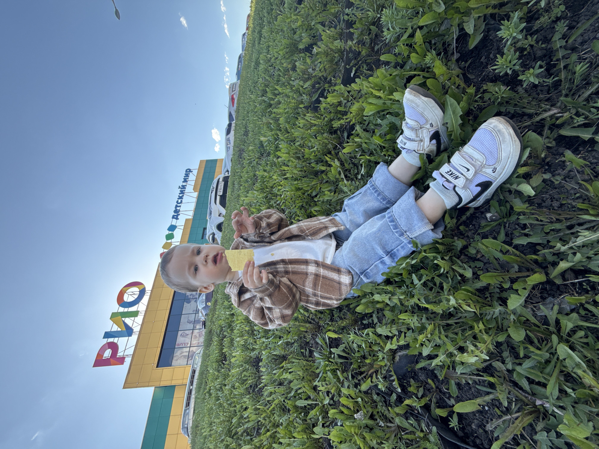 A young child, lying in a field of dandelion weeds near the parking lot of the “RIO” shopping mall in Zavolzhye, Russia. The child is wearing a white t-shirt, jeans, a brown plaid shirt, and dirty white Nike sneakers. The child is looking towards the camera and holding a yellow object, potentially food. The shopping mall is visible in the background, with parked cars in front of it. The sky is blue.