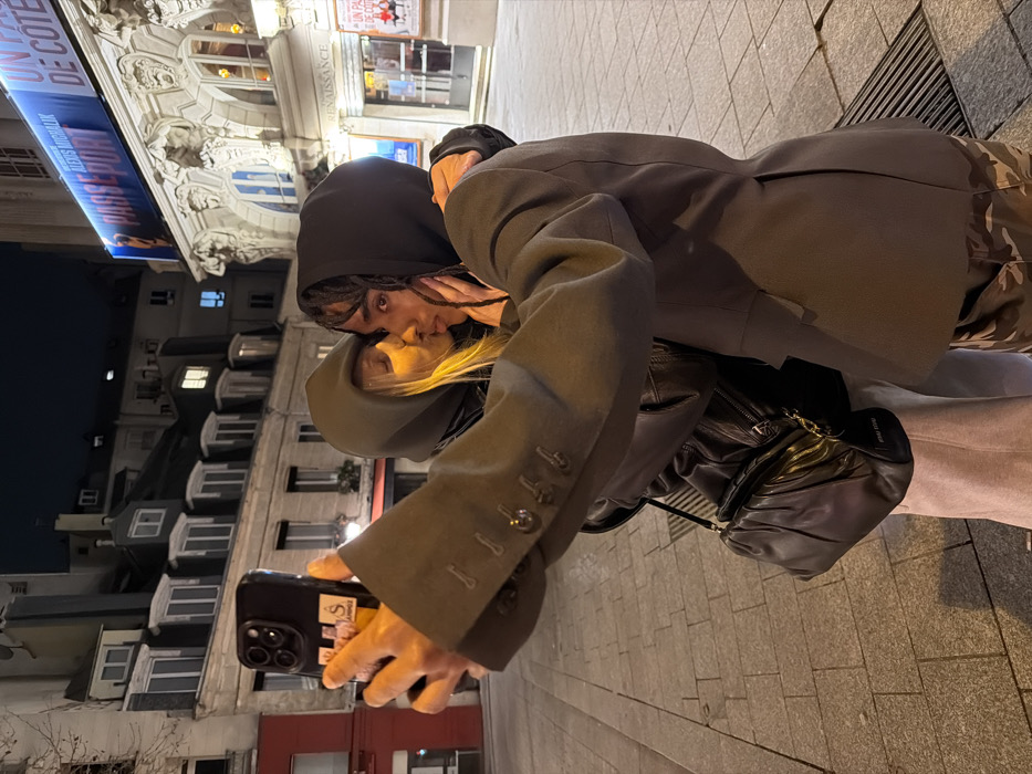 A couple kisses while posing for a phone photo in an urban setting. The person taking the picture wears a gray coat. The kissing couple has one individual wearing a black hooded top, while the other wears a hooded gray jacket. A black purse is slung over the photographer's shoulder. Buildings with windows line the background, and a theater sign is visible at the top. The photo appears to be taken at night.