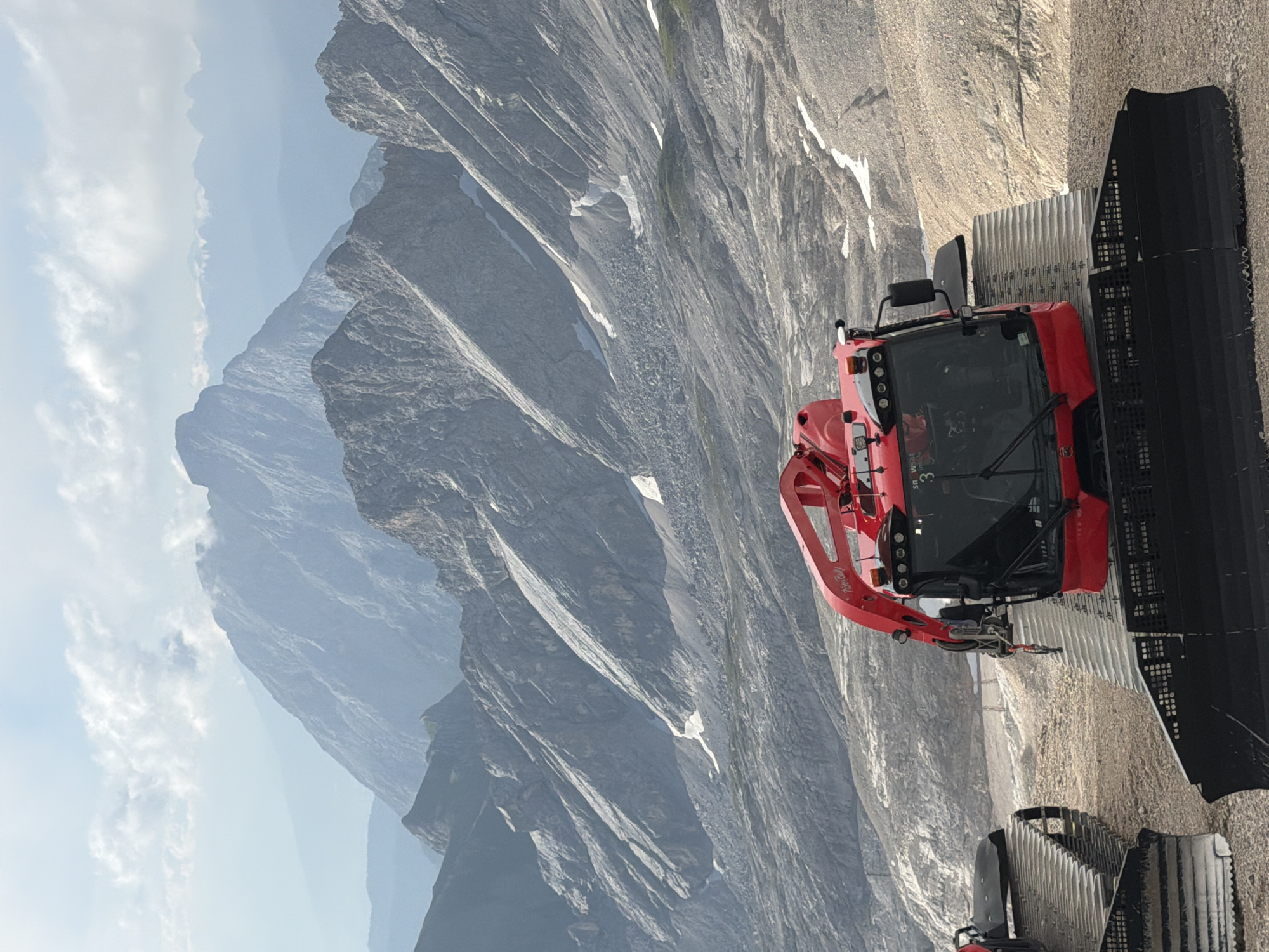 The image shows a red snow groomer in the foreground with a mountainous landscape in the background. The snow groomer is parked on a dirt and gravel surface. The mountains are steep and rocky, with patches of snow visible. The sky is partly cloudy. The image appears to have been taken on a sunny day. The snow groomer has a number "3" on its windshield and writing that says "PistenBully." The location is the Zugspitze, the highest peak in Germany, located near Garmisch-Partenkirchen.