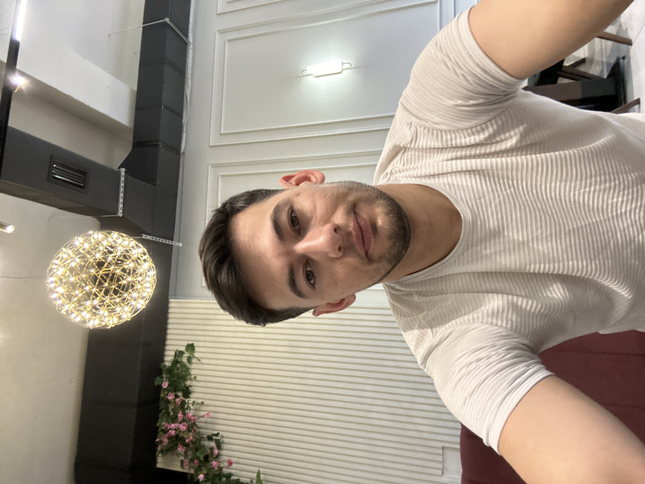 A man in a white, textured shirt is posing for a selfie. He has short, dark hair and a trimmed beard. Behind him is a room with white walls, vertical striped paneling, and ornate molding. A decorative, spherical light fixture hangs to his left, near a black duct and some artificial flowers. A dark chair is partially visible in the top right corner.