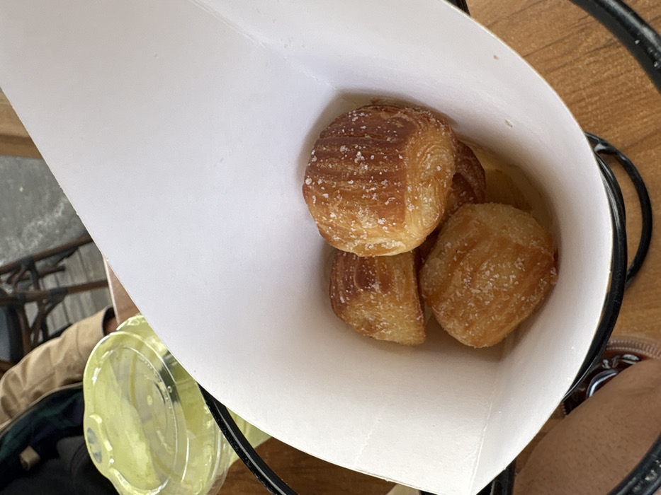 Four pretzel bites coated in coarse salt sit inside a white paper cone holder. The cone is held by a black wire rack on a wooden surface. Part of a clear plastic cup and a person's leg wearing brown shoe are visible. The pretzel bites have a golden-brown color with visible layers.