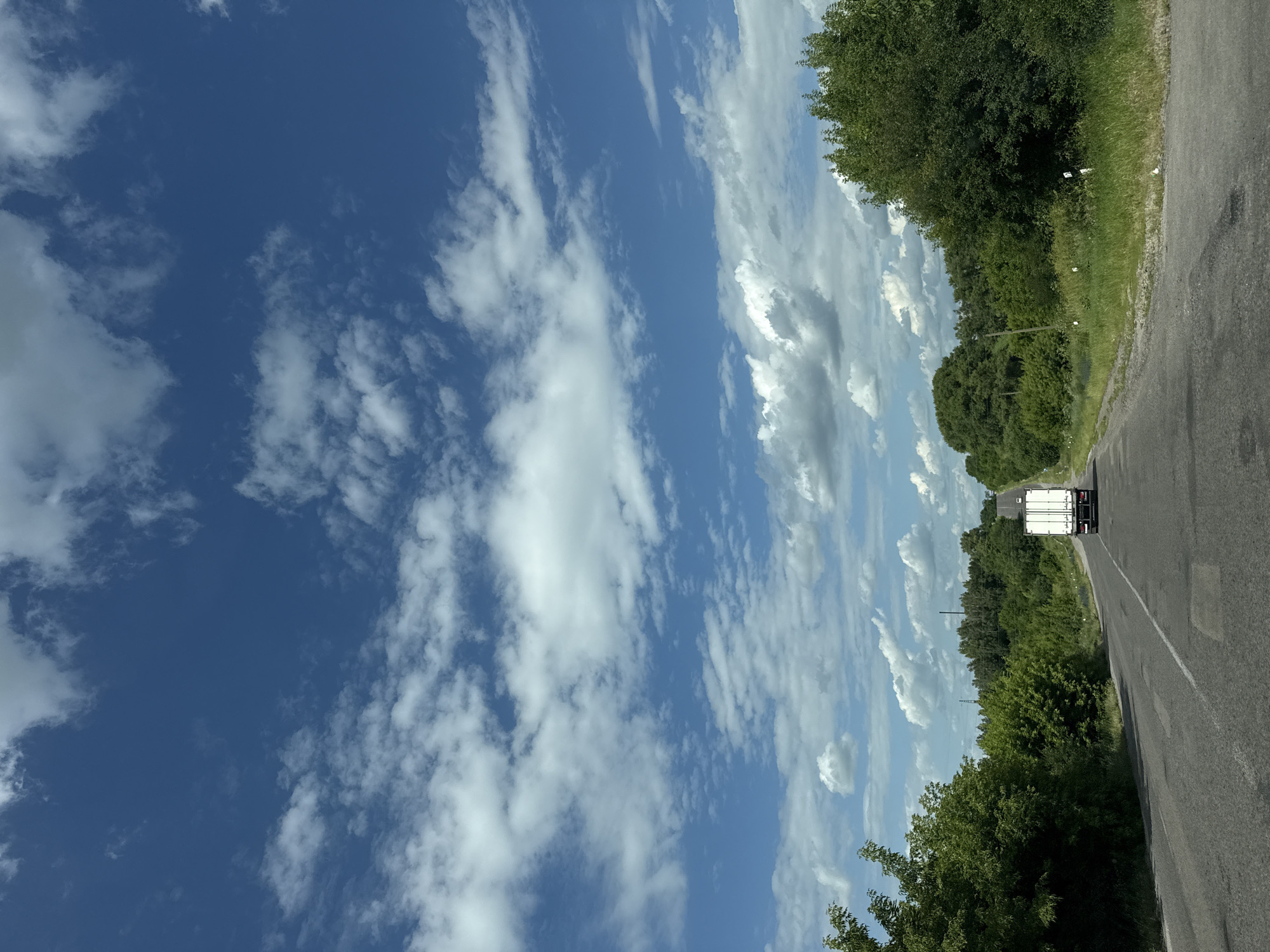 The image depicts a rearview perspective of a two-lane asphalt road winding through a rural landscape under a bright blue sky with scattered white clouds. Dense green foliage lines both sides of the road, with trees and bushes forming a natural canopy. In the distance, a white box truck is traveling away from the viewer on the right lane of the road. The road surface shows signs of wear, with patches and faded lane markings. The overall scene suggests a sunny day in a non-urban environment. The image is not taken within the city of Skvyra, Ukraine.