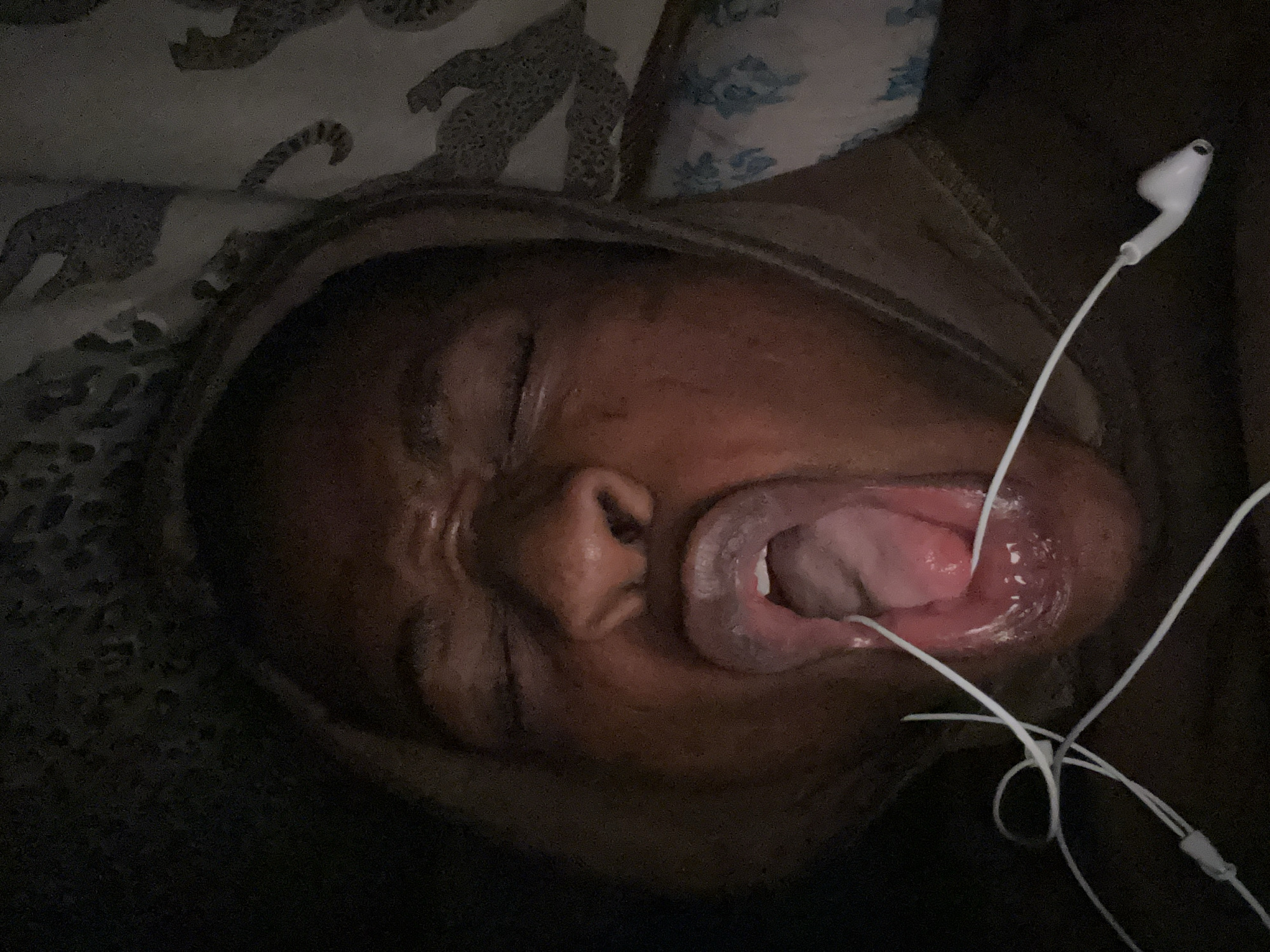 A close-up, dimly lit image depicts a person lying down, likely on a bed, with their mouth open and tongue visible. White earphones are tangled near the mouth. The person's eyes are closed. Bedding with a possible animal print is visible near the head.