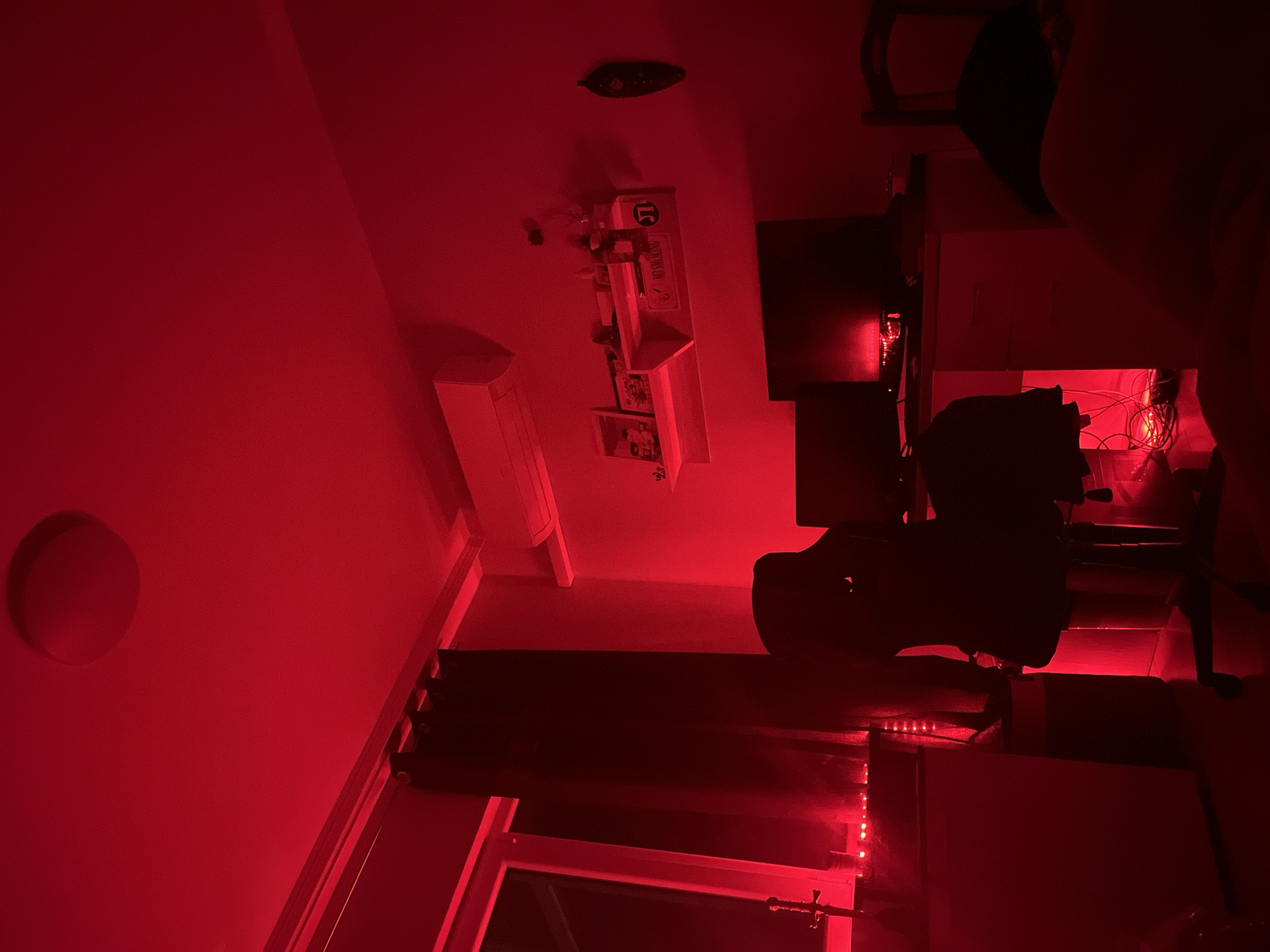 Due to the orientation of the image, it appears to be upside-down.
The scene is dominated by a red light cast across the entire room, making details difficult to discern. The interior setting suggests a study or office space.
Prominent features include:
* A wall with a circular recessed light fixture.
* A wall-mounted shelf cluttered with objects, including a "no smoking" sign and a "15" sign.
* Multiple computer monitors, stacked vertically.
* A desk chair.
* A window covered by dark curtains with red LED lights.
No people, subjects, actions, or interactions are apparent.
It is not possible to locate this scene within Plovdiv, Bulgaria based on this visual information.