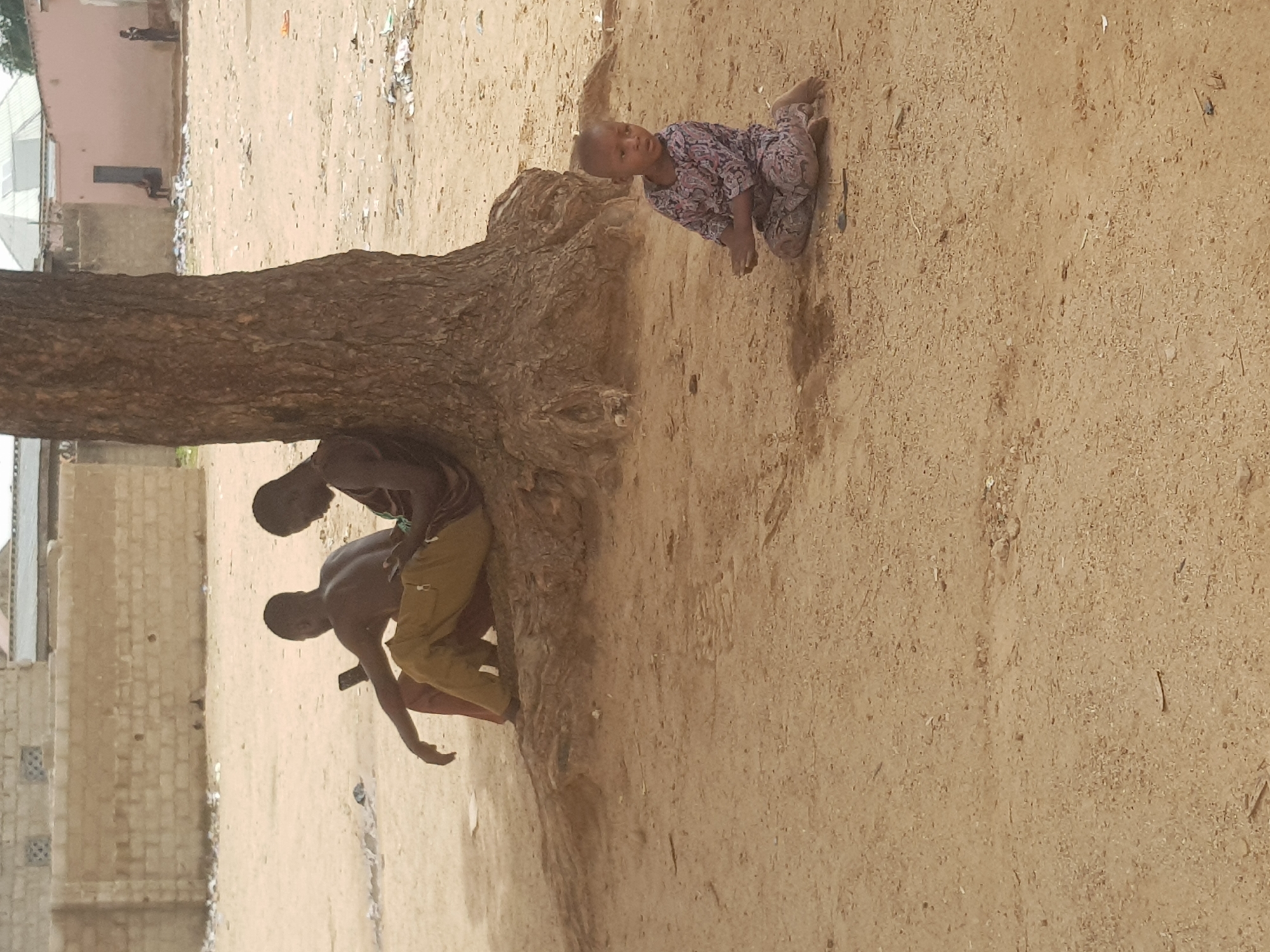 The scene takes place on a sandy ground in what appears to be Dabawa, Nigeria. Two individuals are positioned near the base of a large tree. One person is lying on their stomach, with their body obscured behind the tree trunk and branches. Another individual kneels nearby. A third person, a child wearing patterned clothing, is crawling on the sandy ground away from the tree. In the background, buildings are visible. Litter and debris is scattered on the ground.