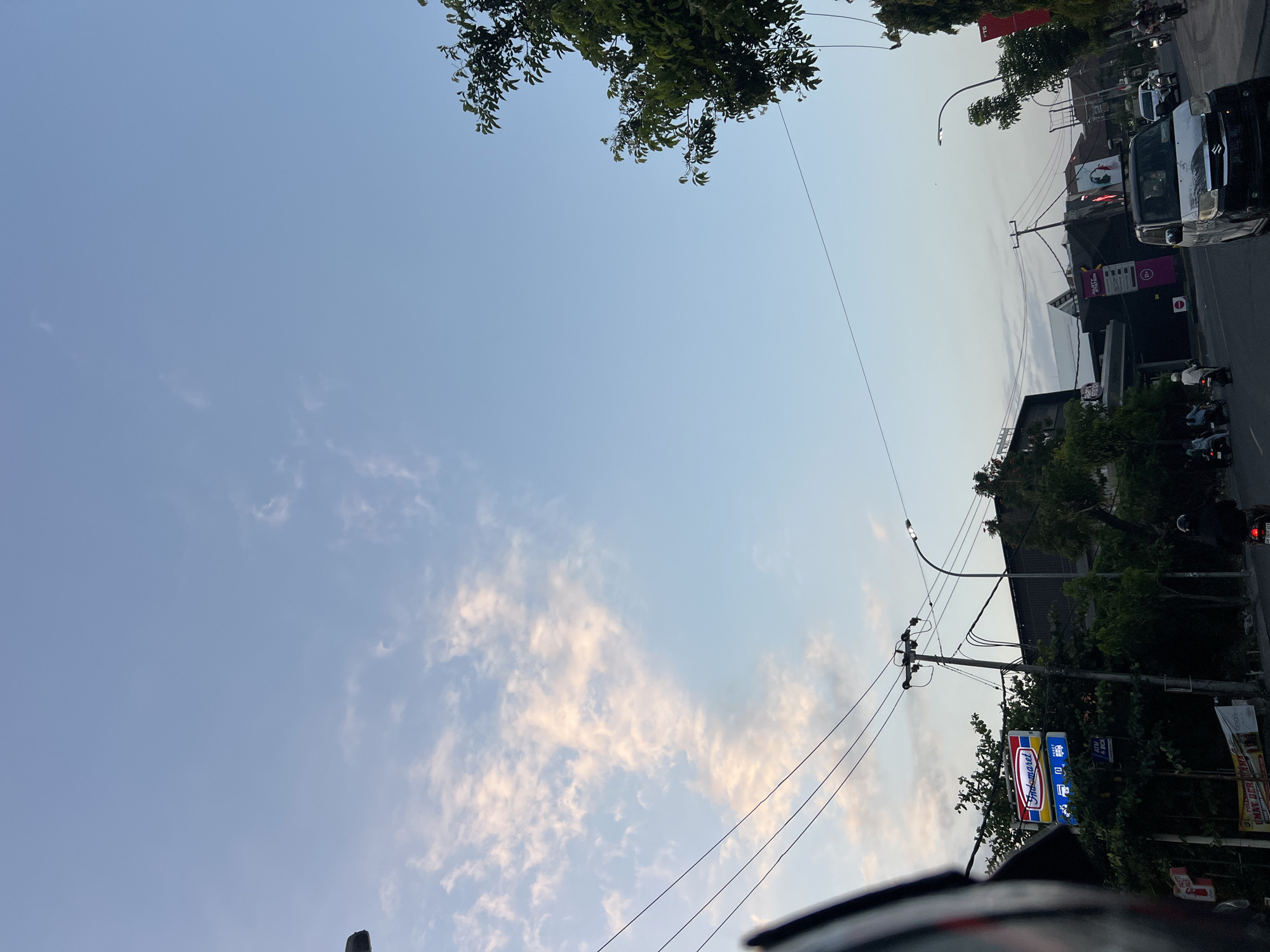 The image depicts a street scene in Kuta, Indonesia, under a pale blue sky with scattered, wispy clouds. In the foreground, a portion of a motorcycle helmet is visible on the left. A prominent Indomaret convenience store sign, featuring its characteristic red, yellow, and blue branding, is located on the left side of the street. Trees line the side of the road, interspersed with streetlights and utility poles with numerous electrical wires. Several people are present on motorcycles and a small white car is driving on the road. The scene appears to be during the day, possibly late afternoon or early evening, given the soft lighting. Buildings and businesses are visible in the background.