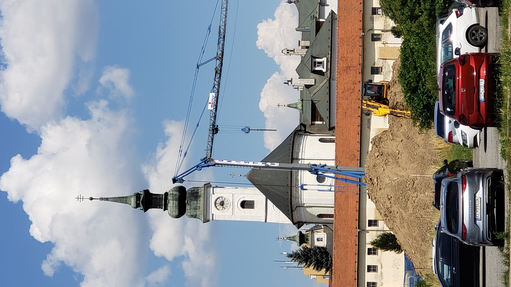 The image depicts a construction scene with a large crane positioned in front of a white church featuring a tall, green-domed steeple with a clock. The church has a dark, conical roof. In the foreground, several cars are parked. Behind the church, a large pile of dirt is visible, with a yellow excavator partially submerged in it. The sky is blue with scattered white clouds. The scene appears to be in the city of Prešov, Slovakia, with the prominent church possibly being the Church of St. Nicholas, a historical landmark in the city. The crane has the logo "UNISTAV" on it, suggesting a construction company is involved in the project.
