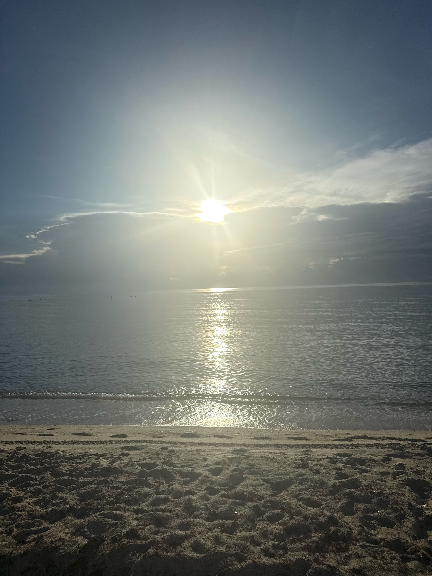 The image depicts a tranquil beach scene in Sunny Isles Beach, United States of America, at sunrise or sunset. The sun, a bright orb, is positioned in the upper center of the frame, partially obscured by clouds. Its rays create a shimmering path of light across the calm ocean surface. Gentle waves lap at the shore, leaving a wet, reflective line on the sandy beach in the foreground. The sand is textured with footprints and subtle undulations. The sky transitions from a pale blue at the top to lighter, hazy tones near the horizon, with scattered clouds catching the sunlight. In the distance, on the water, a few dark specks suggest the presence of distant boats or buoys. The overall atmosphere is serene and peaceful.