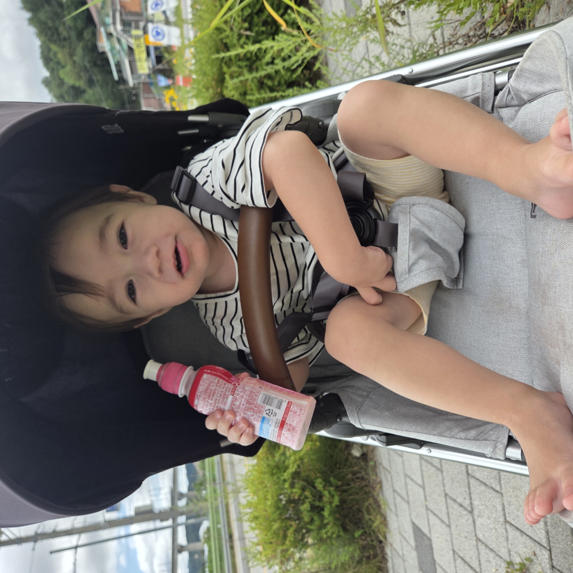 A toddler with light brown hair and brown eyes sits in a stroller, holding a pink bottle of liquid. The toddler wears a striped t-shirt and yellow and white striped shorts. The stroller has a brown handlebar and gray safety straps. The background shows a street with buildings and greenery. This image was taken in Dunnae-myeon, South Korea.