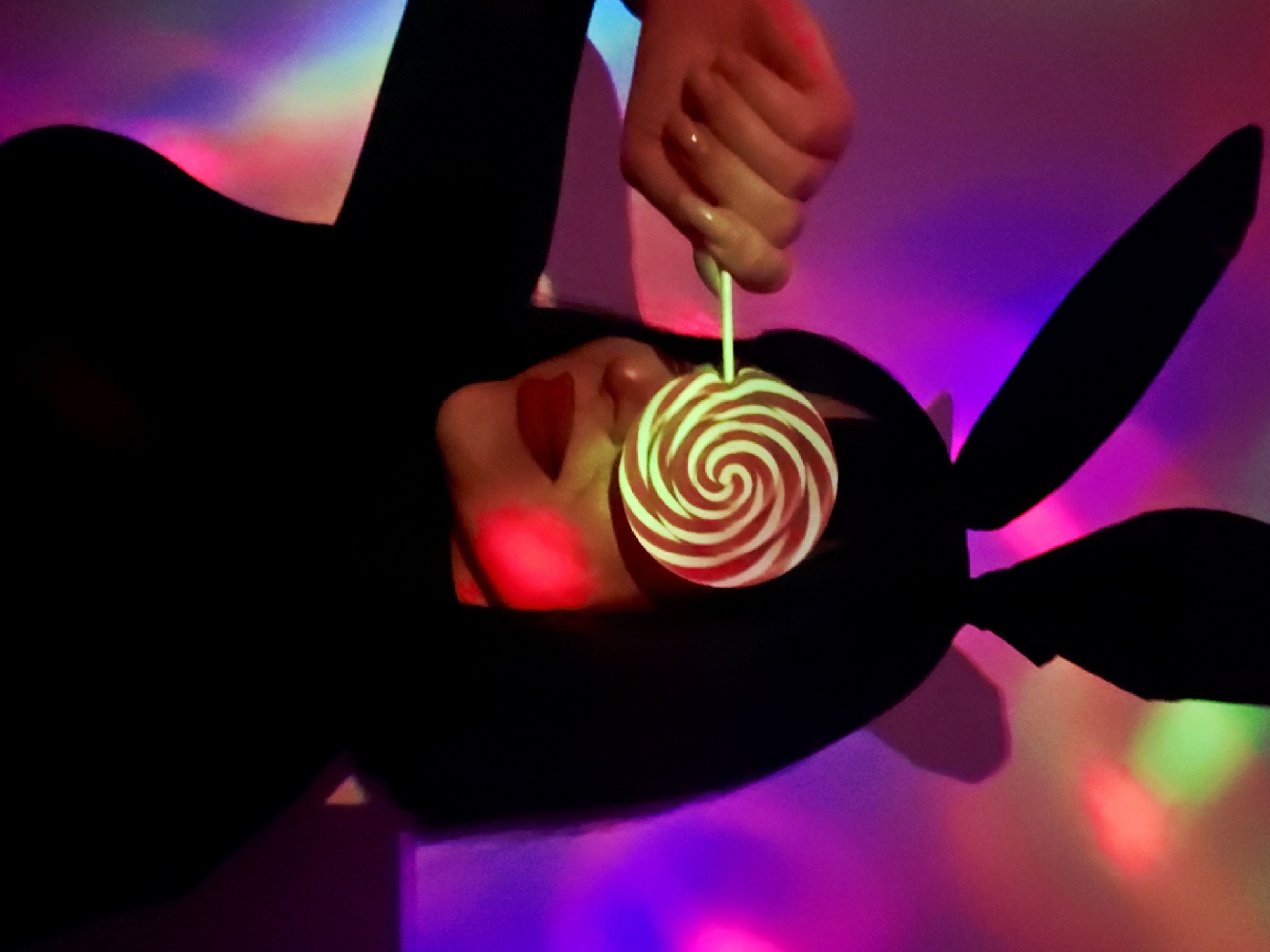 A person lies down, wearing bunny ears and holding a red and white swirly lollipop in their hand, which is positioned near their face. A heart is painted on their cheek. The scene is lit with a variety of neon colors, including pink, purple, green, and red, which reflect off the surface beneath them.