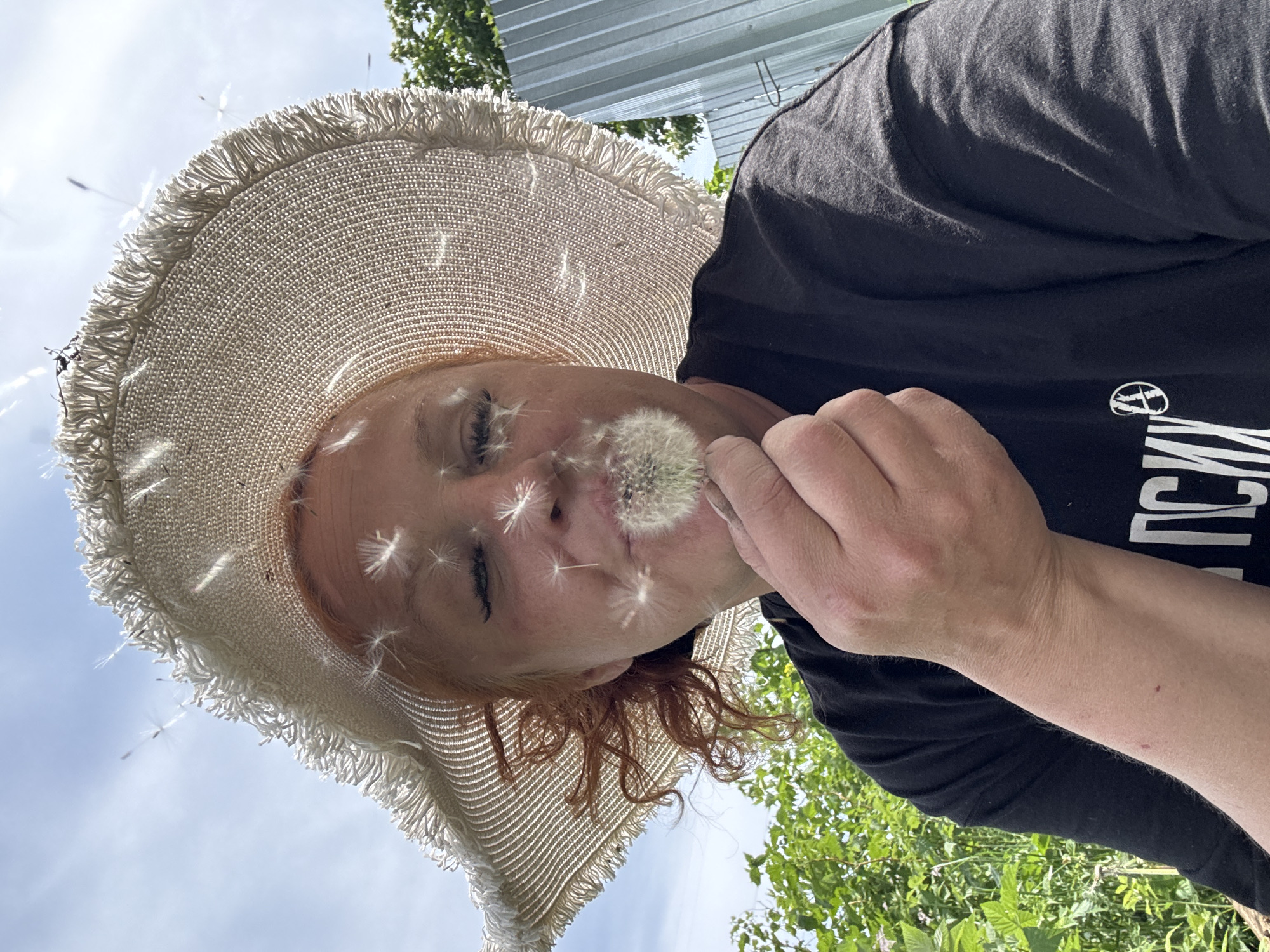 A person wearing a white, wide-brimmed hat and a black t-shirt holds a dandelion close to their face. The person has reddish hair. Dandelion seeds float around their face. The background includes a glimpse of a blue sky, green foliage, and a silver corrugated metal structure. The location is Chaikovsky, Russia.