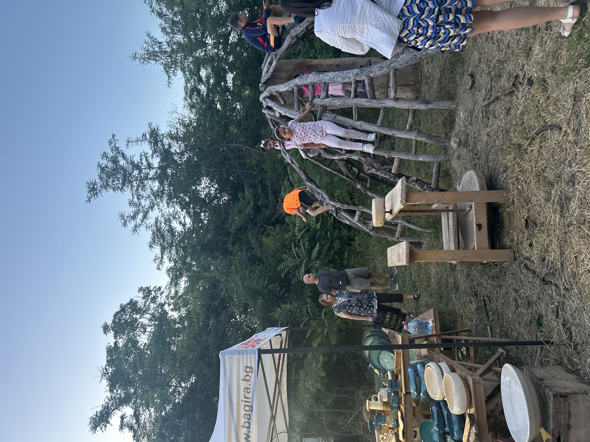 In a rural outdoor setting with a clear sky, a group of people are gathered near a makeshift wooden play structure and a tent displaying pottery. In the foreground, a woman in a white top and a patterned skirt stands facing away from the camera. To her left, a tent with the text "w.bagira.bg" is set up, displaying various ceramic items like bowls and vases on wooden tables. A bottle of water is visible on one of the tables. In the mid-ground, the wooden play structure is the focal point, with several children climbing on it. A boy in an orange shirt is on the left side of the structure, while a girl in a pink and white spotted shirt is climbing a ladder in the center. Another child in pink is visible further up the structure, and a boy in a blue and red shirt is sitting on the top. Adults are also present, with a man and a woman standing near the tent. The ground is covered in dry grass and twigs, with trees forming a dense backdrop.