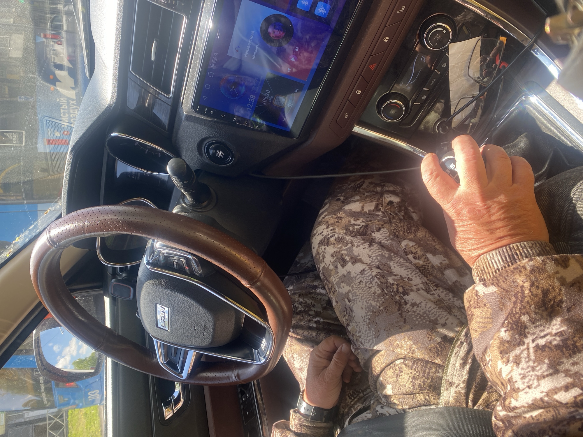The image shows the interior of a car. The person driving is wearing camouflage patterned clothing. The steering wheel is brown leather with a silver center where the brand logo "LIFAN" is displayed. The dashboard features a screen that displays a clock reading "12:38", frequency "106/20 MHz", and Russian text "Неизвестный" ("Unknown"). The driver's hand is on the gear shift. In the bottom left corner of the image, partially visible in the side mirror, there is a blue vehicle with "103" written on it, and the Russian text "Кузбасс Чистый Воздух" ("Kuzbass Clean Air"). The presence of this text suggests that this is Novokuznetsk, Russia, due to the prominence of the Kuzbass region.