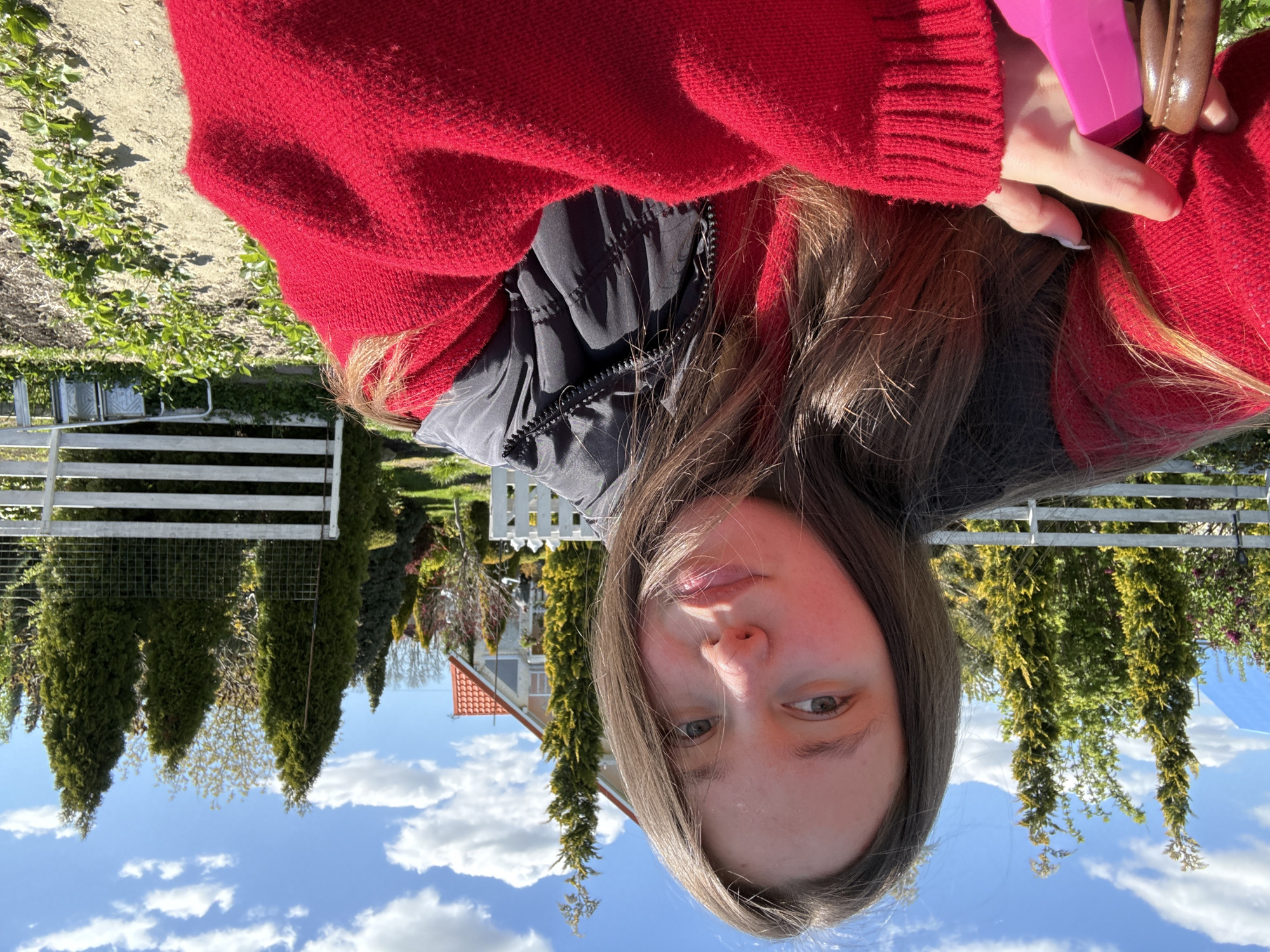 A portrait-style photograph captures a young individual outdoors in what appears to be a garden or yard in Pervorechenskoye, Russia. The image is taken from a slightly low angle, looking up towards the subject.
The subject is a young person, likely a female teenager or young adult, with long, light brown hair partially covering her shoulders and flowing around her face. She is wearing a bright red knitted sweater or cardigan over a black puffer jacket or vest. Her right hand is visible, holding a vivid pink object, possibly a smartphone or a small container, and a brown, possibly wooden, bracelet or hair tie is on her wrist. She looks directly towards the camera with a calm, neutral expression.
The background consists of a bright blue sky with scattered white cumulus clouds, suggesting a sunny day. Below the horizon line, a well-maintained outdoor environment is visible. A white wooden picket fence runs horizontally across the middle ground. Behind the fence are several tall, slender evergreen trees (conifers) and other green foliage. In the distance, part of a house with a distinct orange or red tiled roof can be seen amidst more greenery. A patch of cultivated ground with small green plants is visible in the lower left portion of the background. The lighting indicates it is daytime, likely mid-morning or afternoon, with clear weather. No visible text is present in the image.