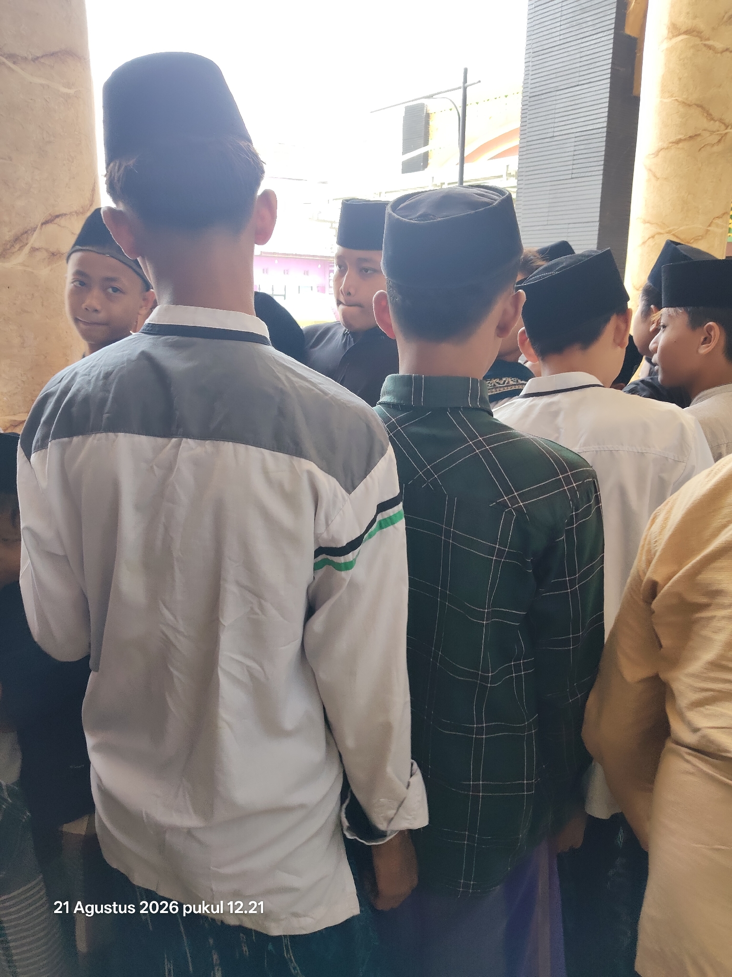The image shows a group of young males wearing traditional Islamic skullcaps and attire, standing in what appears to be an outdoor or semi-outdoor location. The foreground is dominated by the back of two individuals. The person on the left is wearing a light-colored jacket with grey paneling across the shoulders and chest, and green and black stripes on the sleeve. The person to their right is wearing a dark green plaid shirt. Several other individuals are visible in the background, also wearing similar skullcaps. The environment features architectural elements, possibly columns or pillars, with a textured, beige-colored surface on the right and a lighter, possibly stone column on the left. Behind them, there's a building with grey siding and a glimpse of pink structures, suggesting an urban or semi-urban setting. A date stamp in the lower left corner reads "21 Agustus 2026 pukul 12.21". Based on the provided information, this scene is located in Kalitidu, Indonesia.