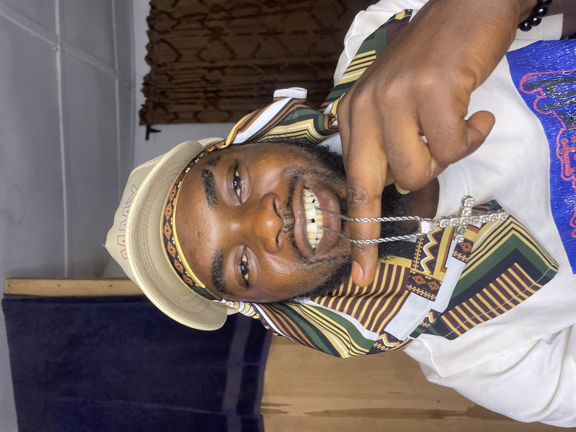 A man with dark skin and a beard is shown in a close-up portrait. He is smiling, and he appears to be holding a silver cross necklace near his mouth. He is wearing a hat with a patterned brim and a patterned scarf around his head and neck. He is also wearing a white shirt with a blue design on it.
The background includes a dark blue surface and a textured brown wall. Based on information inferred from the image's metadata, the photo was taken in the city of Jos, Nigeria.