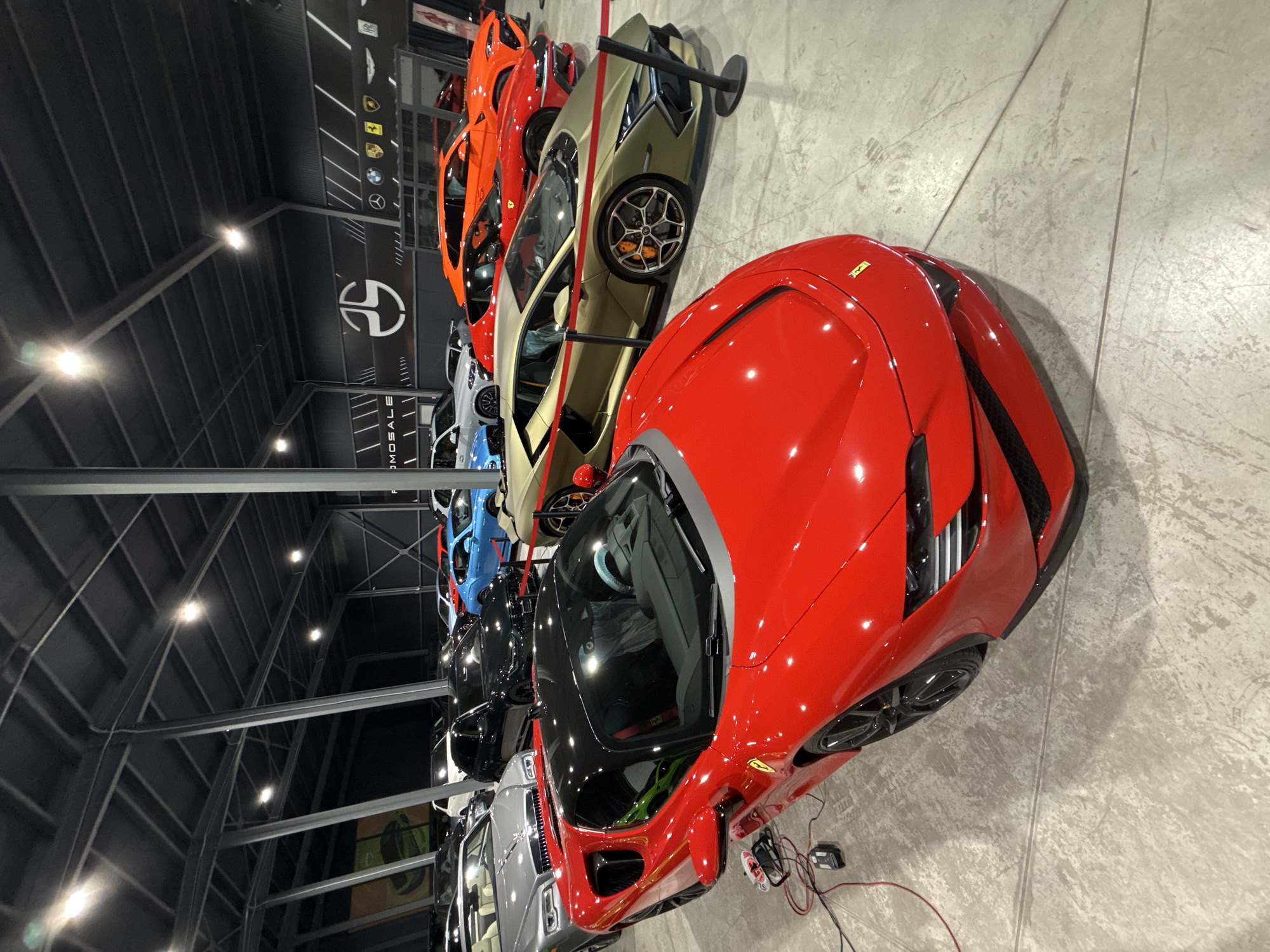 The image depicts an indoor showroom filled with high-end vehicles. In the foreground, a bright red sports car, likely a Ferrari, is prominently displayed. Its sleek design and glossy finish are emphasized by the ambient lighting. Behind it, several other luxury cars are parked in rows, including a gold Lamborghini, an orange Lamborghini Urus, and a blue sports car. The showroom features a modern industrial design with exposed metal beams and a dark ceiling. The floor is a polished concrete surface. A large banner with various car brand logos, including BMW, Mercedes-Benz, and Porsche, is visible on the back wall, along with a stylized logo that appears to be a circular design. The overall impression is one of a well-organized and upscale automotive dealership or private collection. The specific location of this showroom in Plovdiv, Bulgaria is not evident from the image itself.
