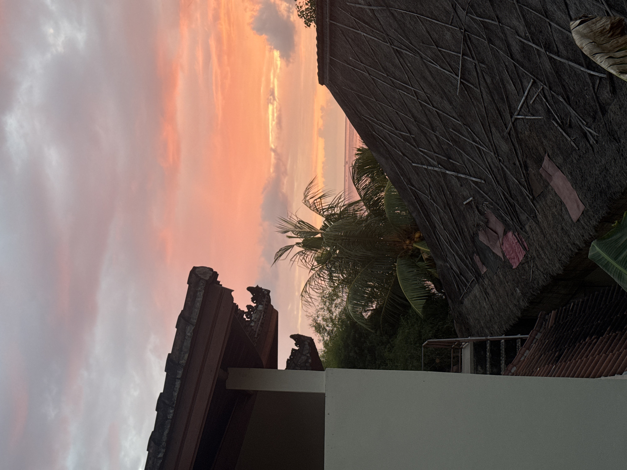 This image captures a sunset view in Abang, Indonesia, with a focus on traditional roofing and tropical vegetation against a vibrant sky. In the foreground, the left side of the frame features the side of a light-colored building with a dark tiled roof. Decorative carvings are visible under the overhang of this building's roof. To the right, a traditional thatched roof with visible wooden support beams dominates the mid-ground, showing signs of wear and repair with patches of dark material. Palm fronds from a coconut palm tree are prominently displayed on the right side of the thatched roof, with coconuts visible. Behind the thatched roof, a dense area of green foliage, likely bamboo and other tropical plants, is visible. The background reveals a body of water, possibly the ocean, under a sky ablaze with hues of pink, orange, and soft gray clouds, indicative of sunset. A sliver of a distant horizon with a few faint boat silhouettes can be seen where the sky meets the water. A metal staircase is attached to the side of the building on the left, leading down towards the lower tiled roof.